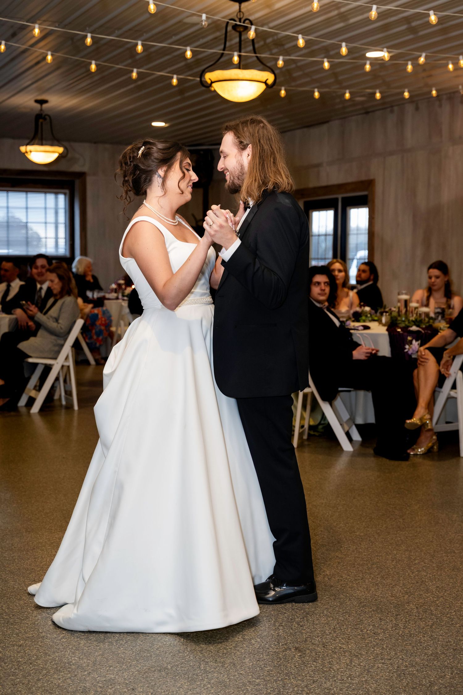 Smiling bride and groom share joyful first dance under warm string lights at wedding reception.