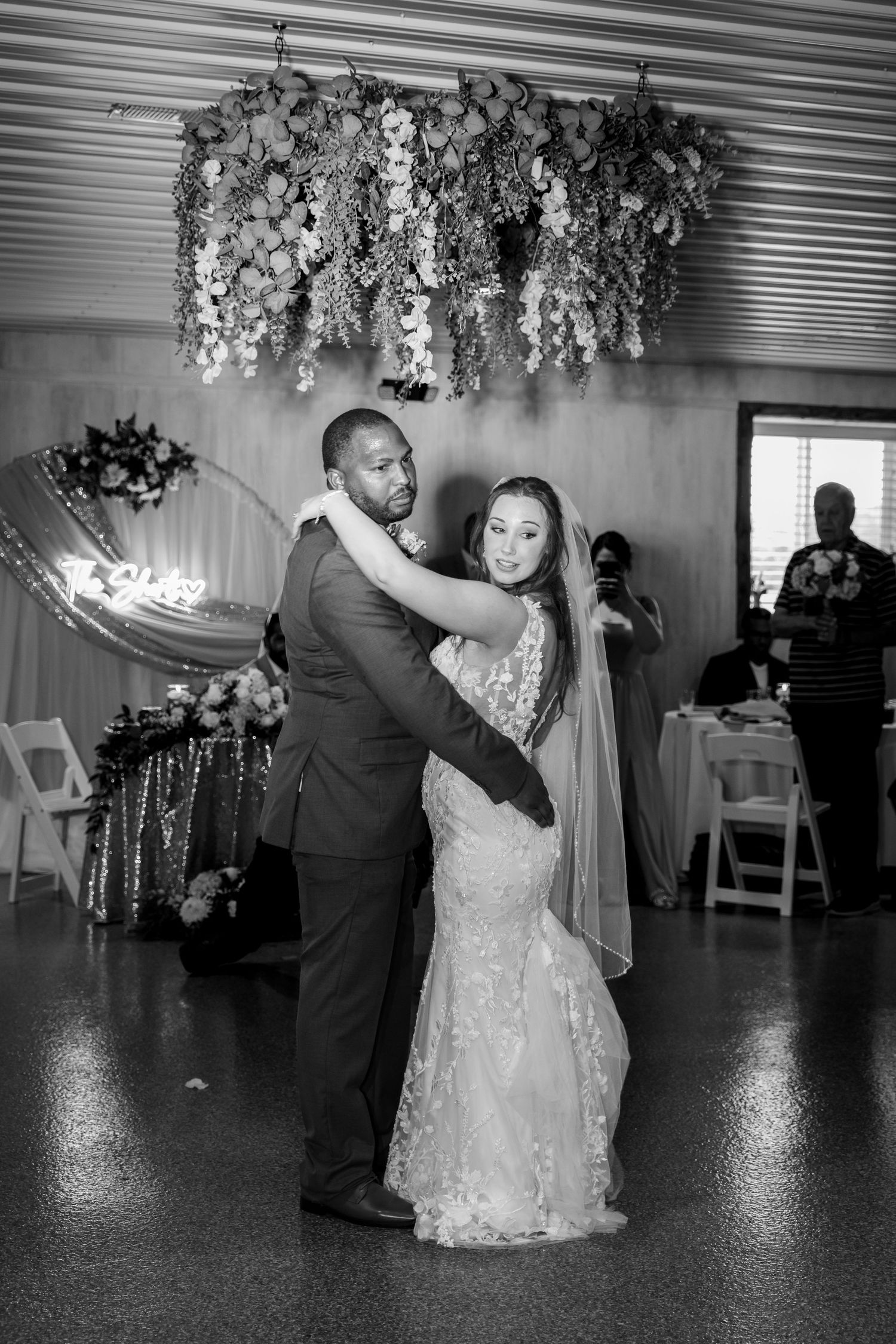 Black and white photo of newlyweds sharing first dance under floral chandelier at elegant wedding reception.
