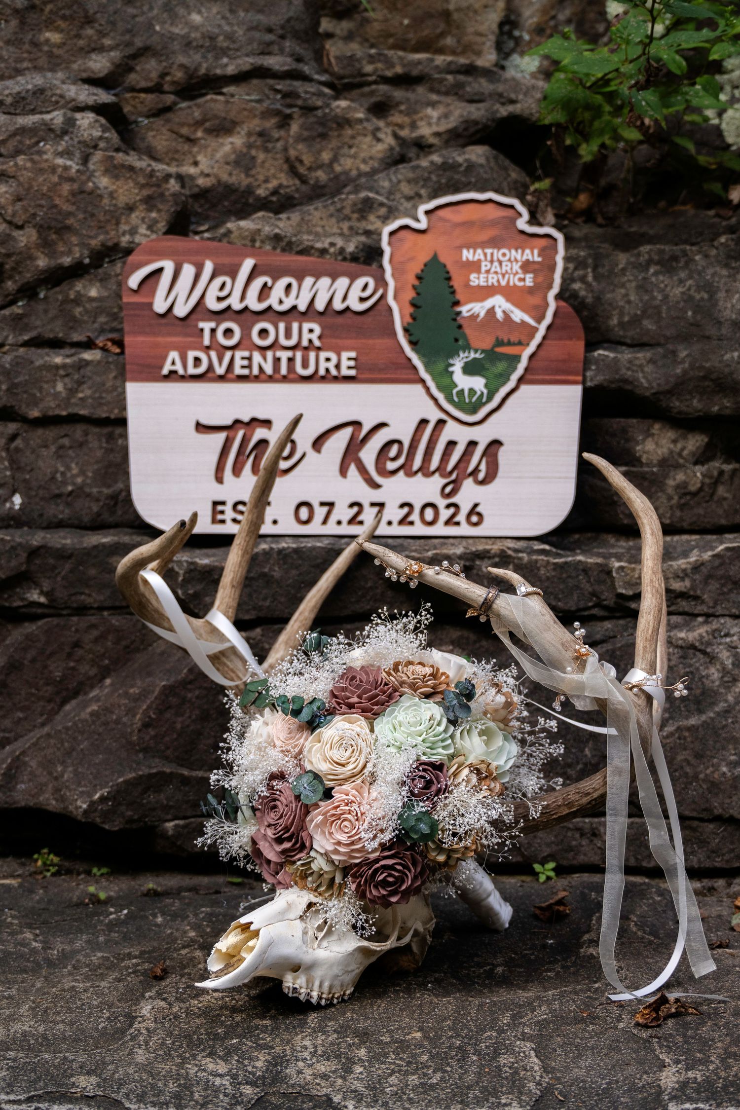 Rustic National Park Service wedding welcome sign for The Kellys with antler and floral bouquet decor on stone wall.