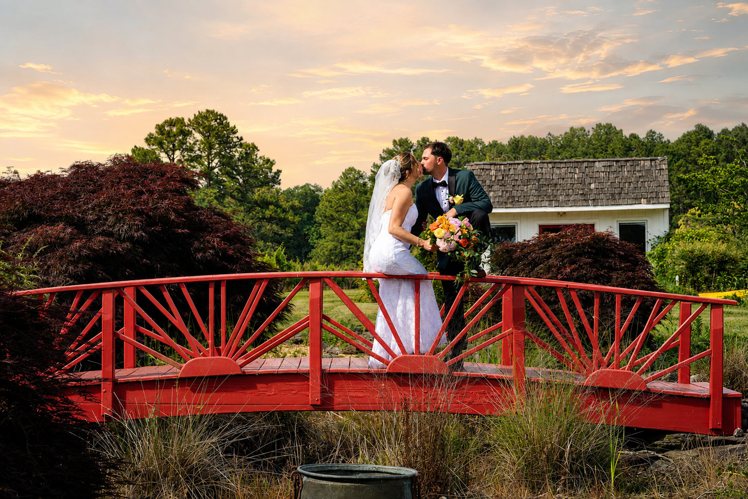 Bride and groom kissing on a red bridge at sunset, surrounded by lush gardens and colorful flowers.