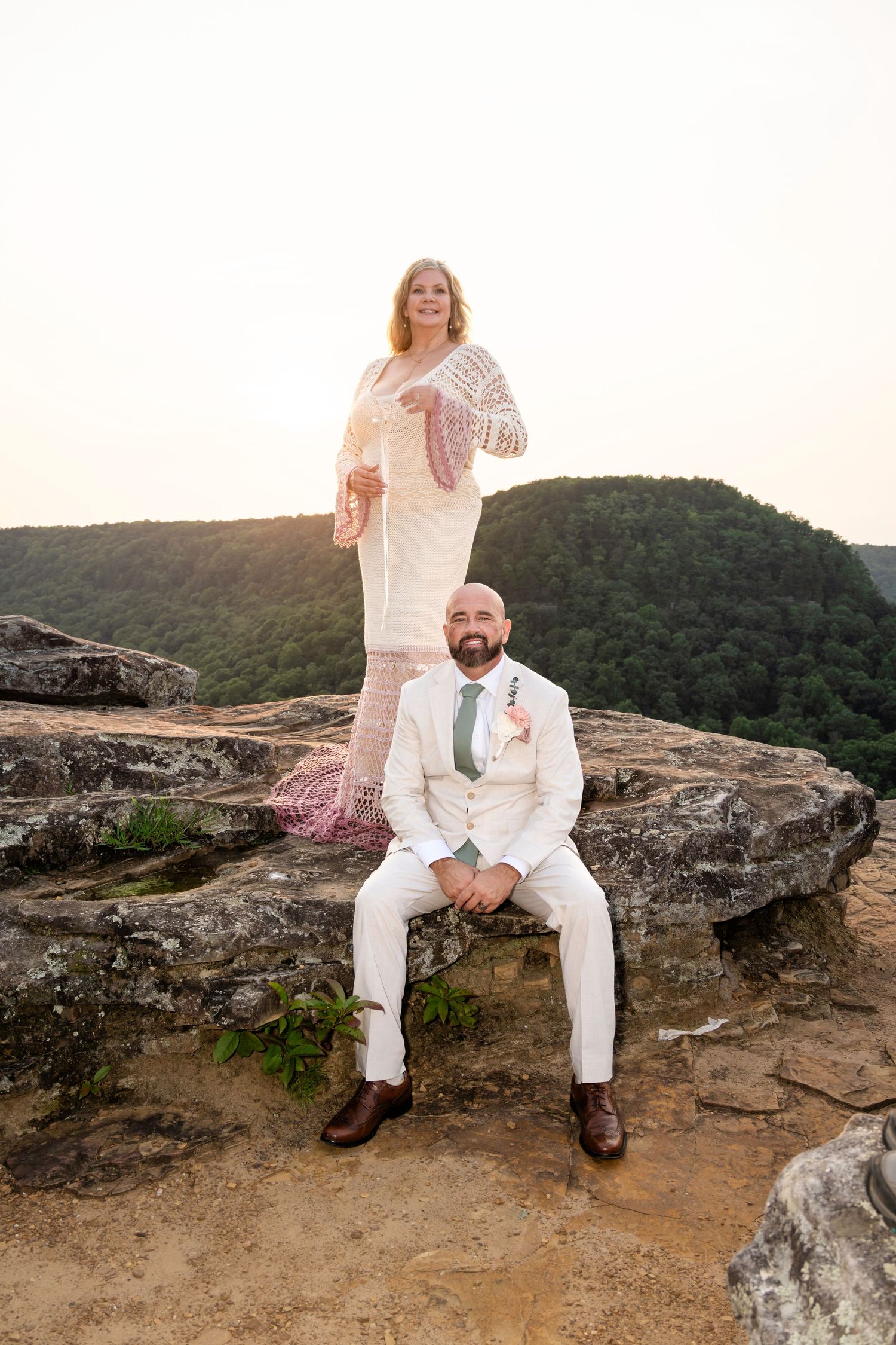 Bride and groom pose on rocky mountain overlook at sunset, she stands behind seated groom in lace dress.