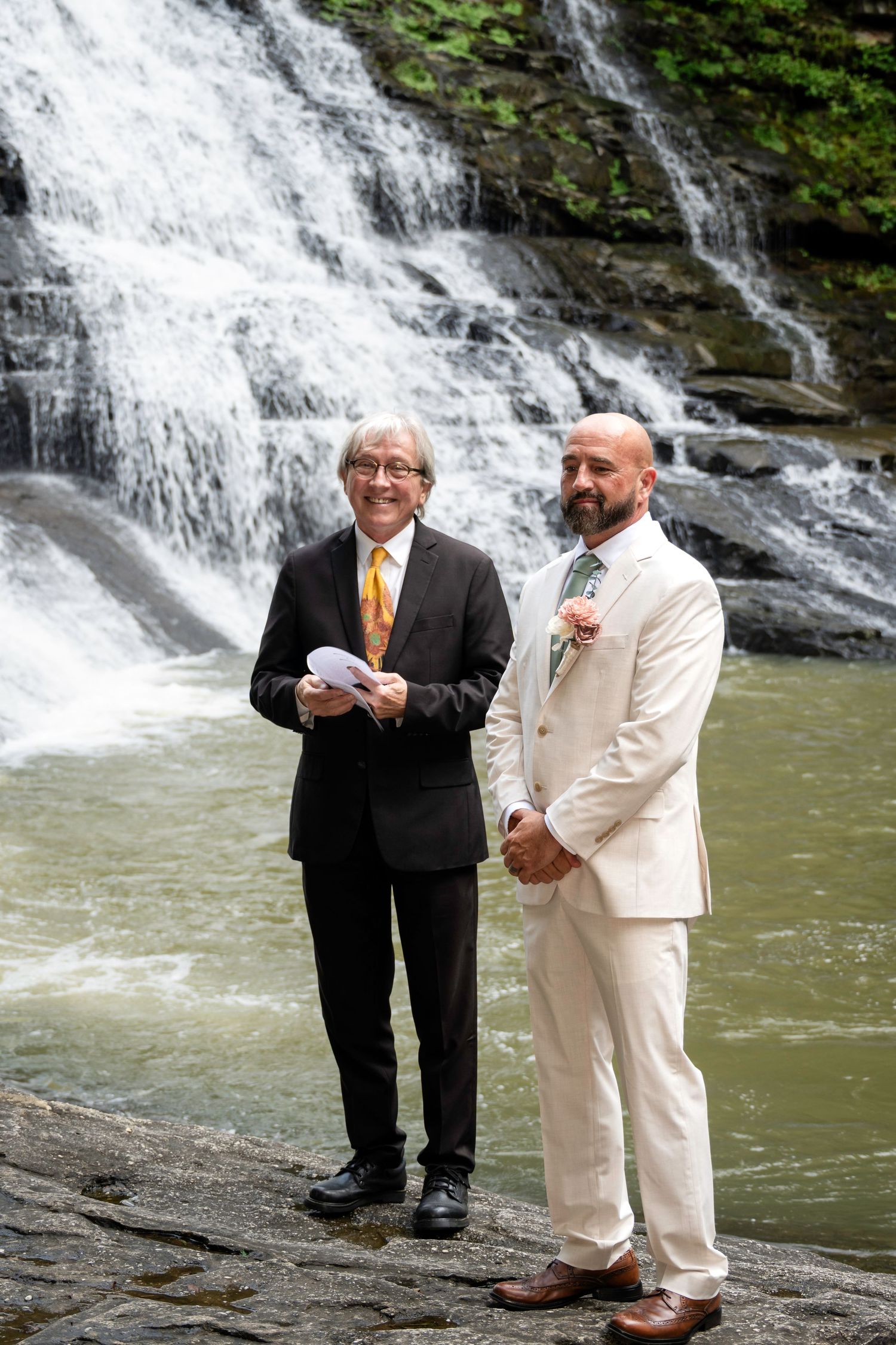 Two men in formal attire stand before a waterfall during an outdoor wedding ceremony.