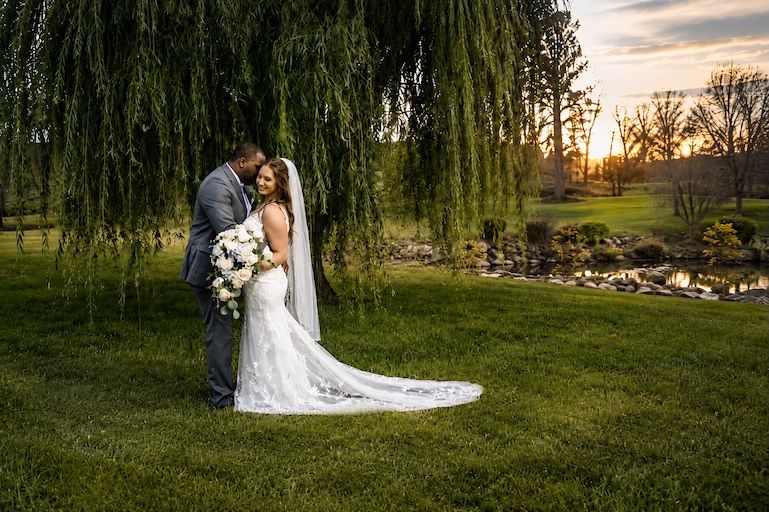 Bride and groom embrace under a weeping willow tree at sunset, with a serene pond and lush green lawn backdrop.