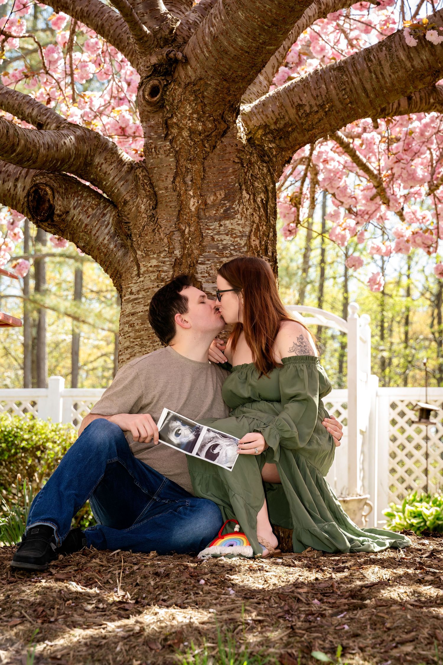 Couple kissing under blooming cherry blossom tree holding ultrasound photos, pregnancy announcement in spring garden.