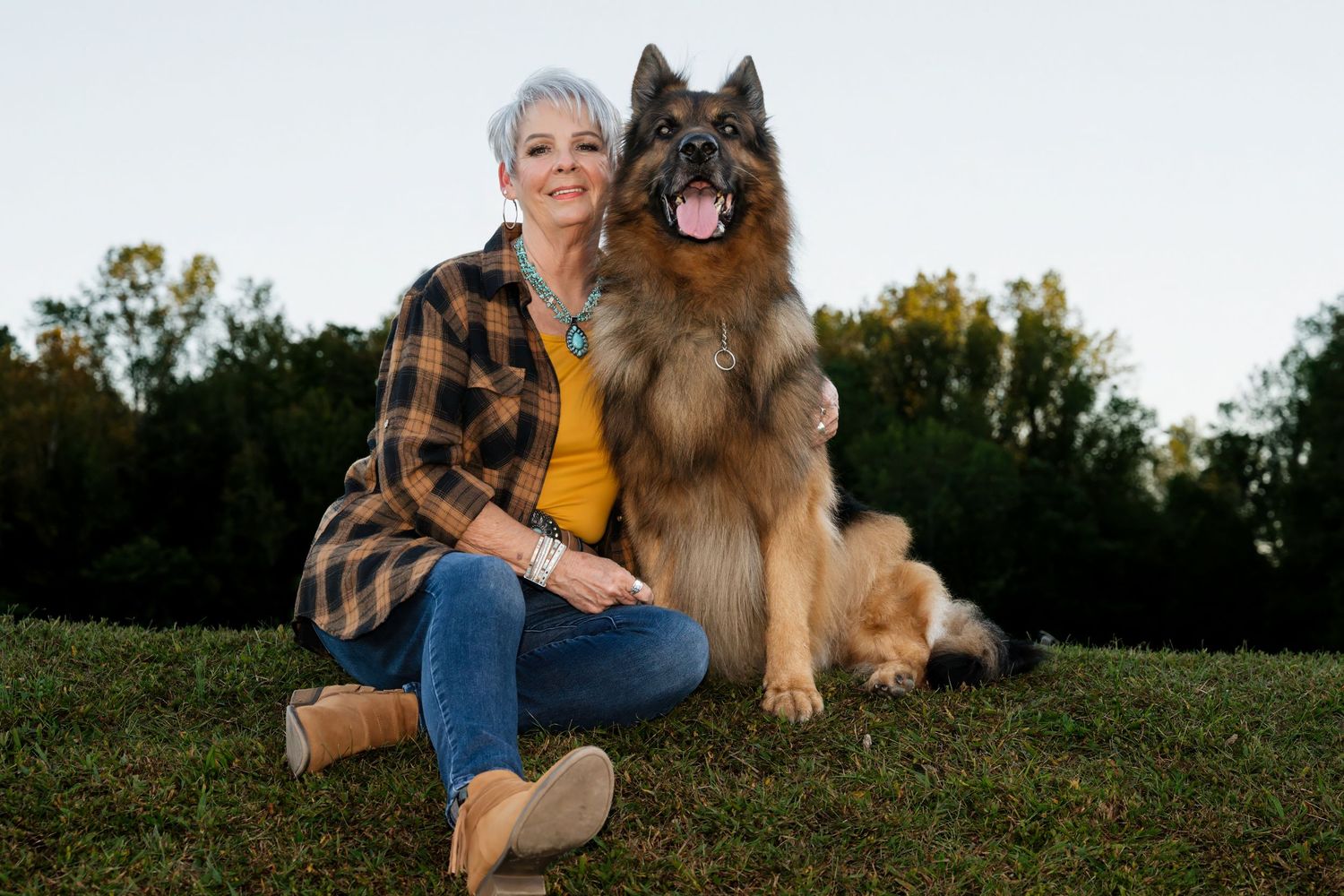 Woman with short gray hair sits on grass hugging a large fluffy German Shepherd dog outdoors.