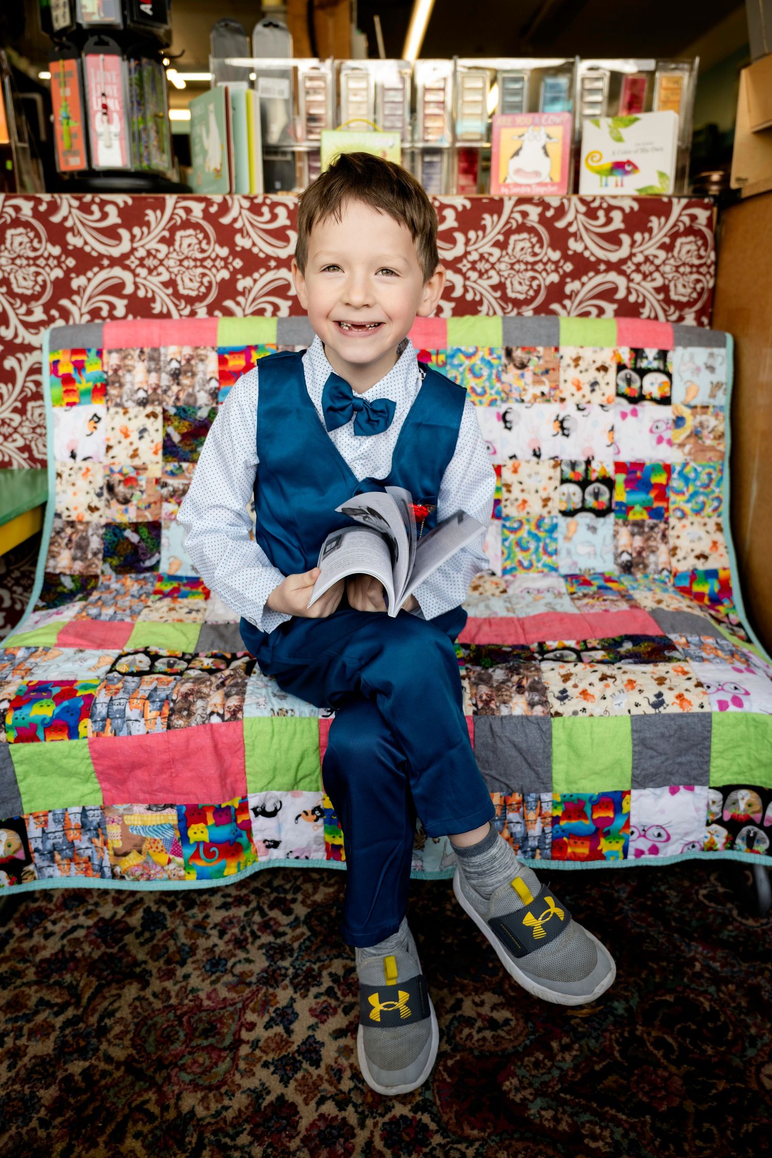 A smiling young boy in a blue vest and bow tie sits on a colorful patchwork quilt, holding a book.