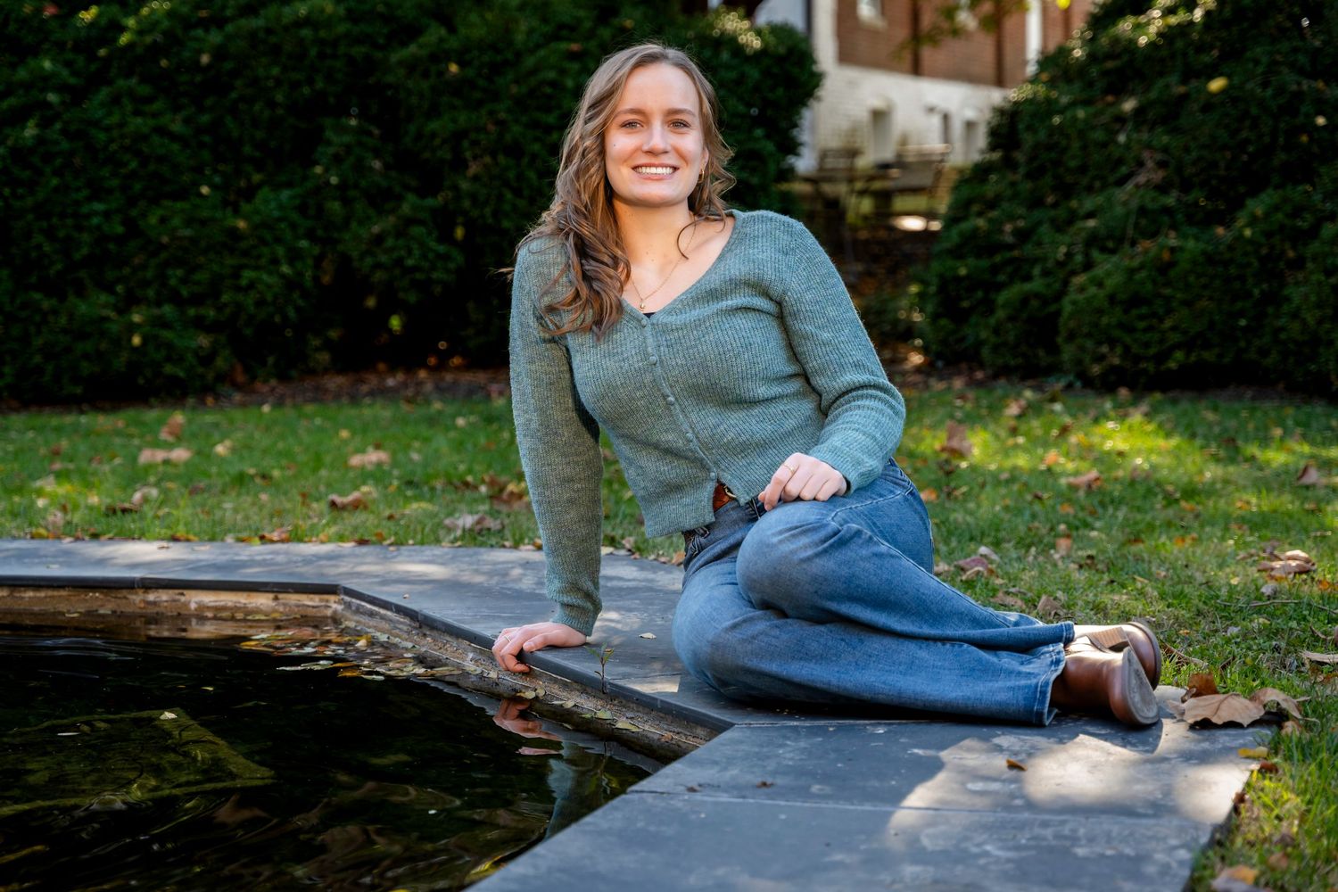 Young woman in green sweater and jeans sitting beside a garden pond smiling outdoors.