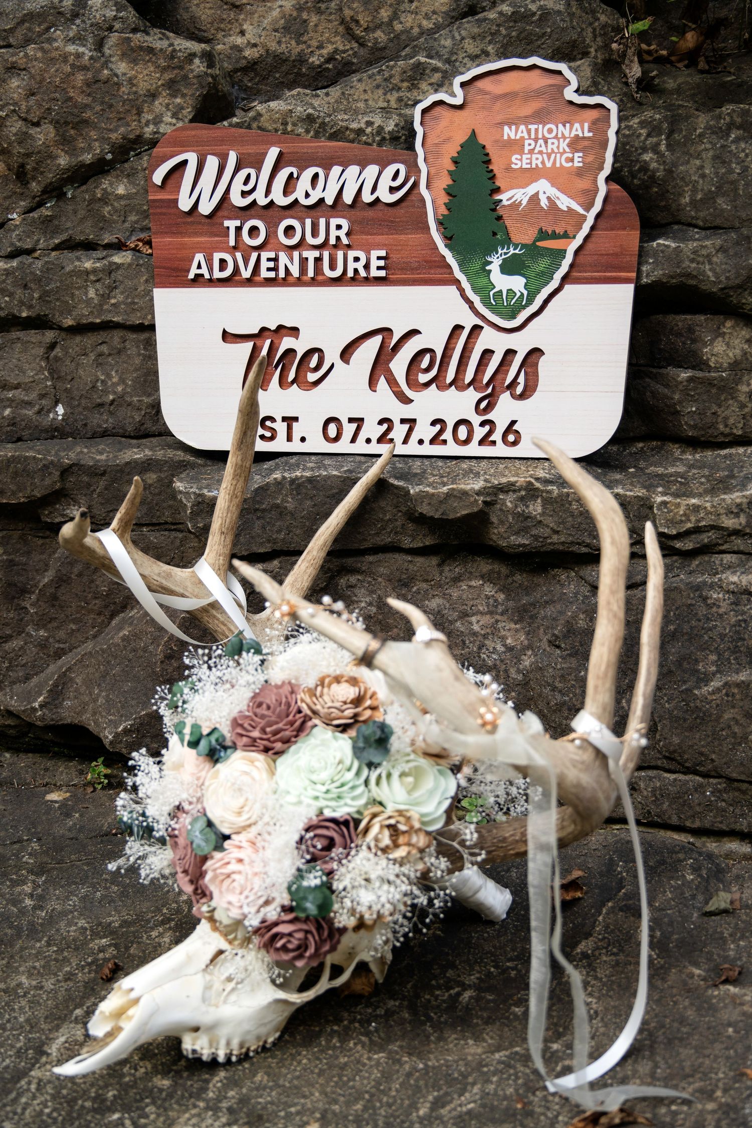 Rustic wedding welcome sign for The Kellys EST. 07.27.2026 with National Park Service theme, deer skull, and floral bouquet.
