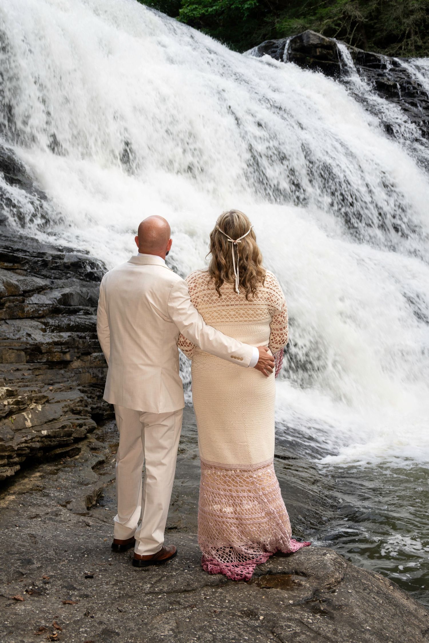 Bride and groom in white standing arm in arm on rocks before a rushing waterfall during an outdoor wedding ceremony.