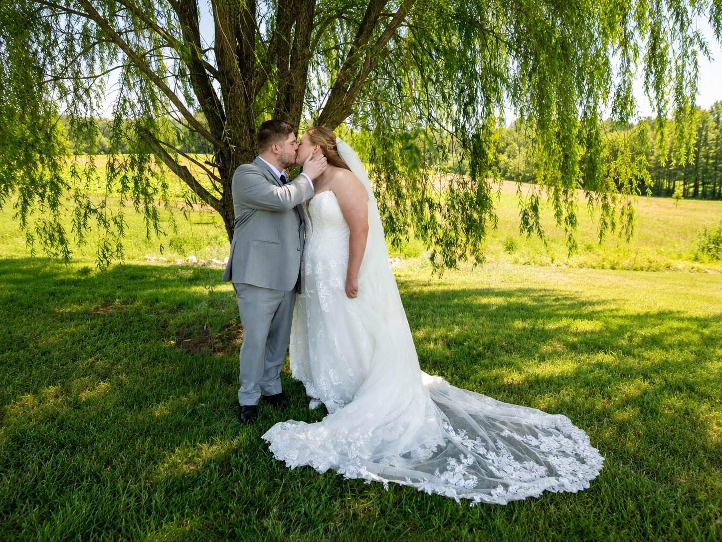 Bride and groom share a romantic kiss under a weeping willow tree on lush green grass during outdoor wedding.