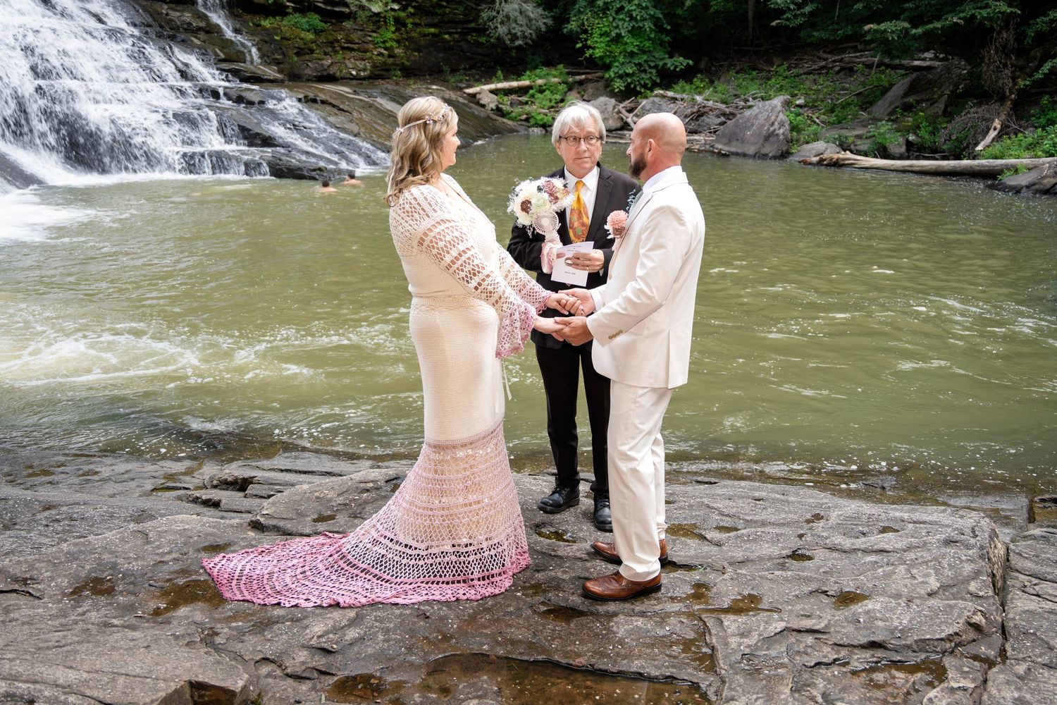Couple exchanging vows with officiant by a waterfall on rocky terrain, bride in pink lace gown, groom in white suit.