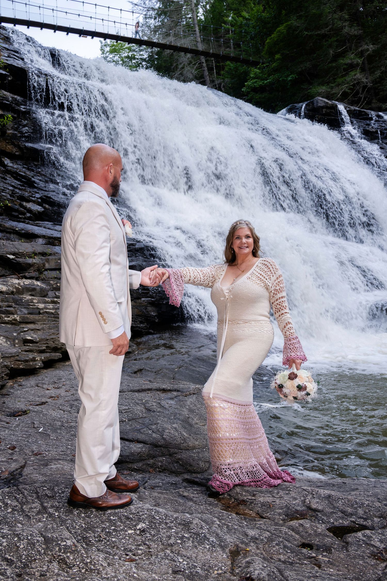 Bride and groom hold hands before a cascading waterfall, she in a lace gown, he in a white suit.