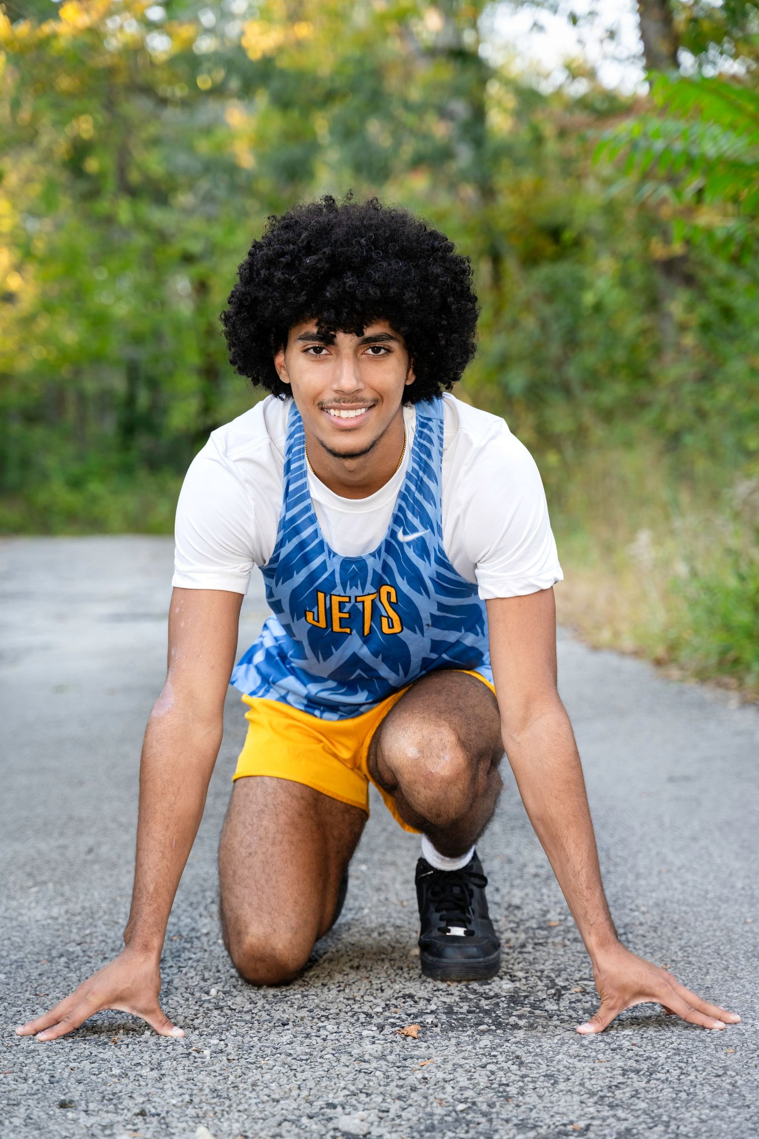 Young male athlete in Jets jersey crouches in starting position on road, smiling confidently outdoors.