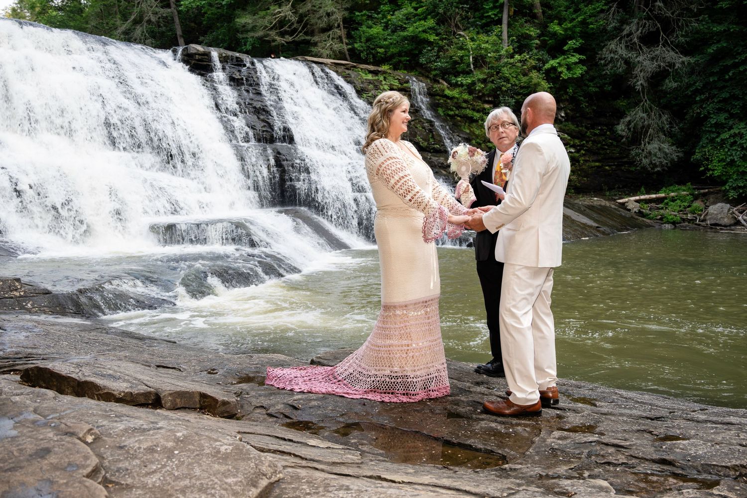 Couple exchanges vows before a stunning waterfall with an officiant on rocky terrain in a lush forest setting.