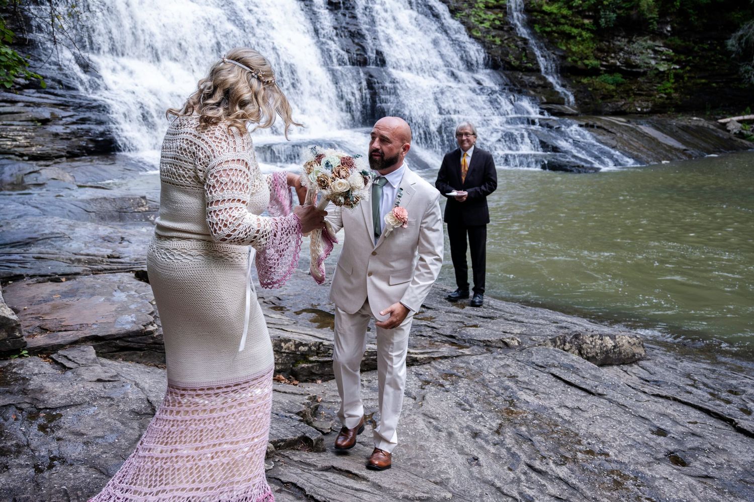 Couple exchanging vows at waterfall wedding ceremony on rocky terrain with officiant in background.