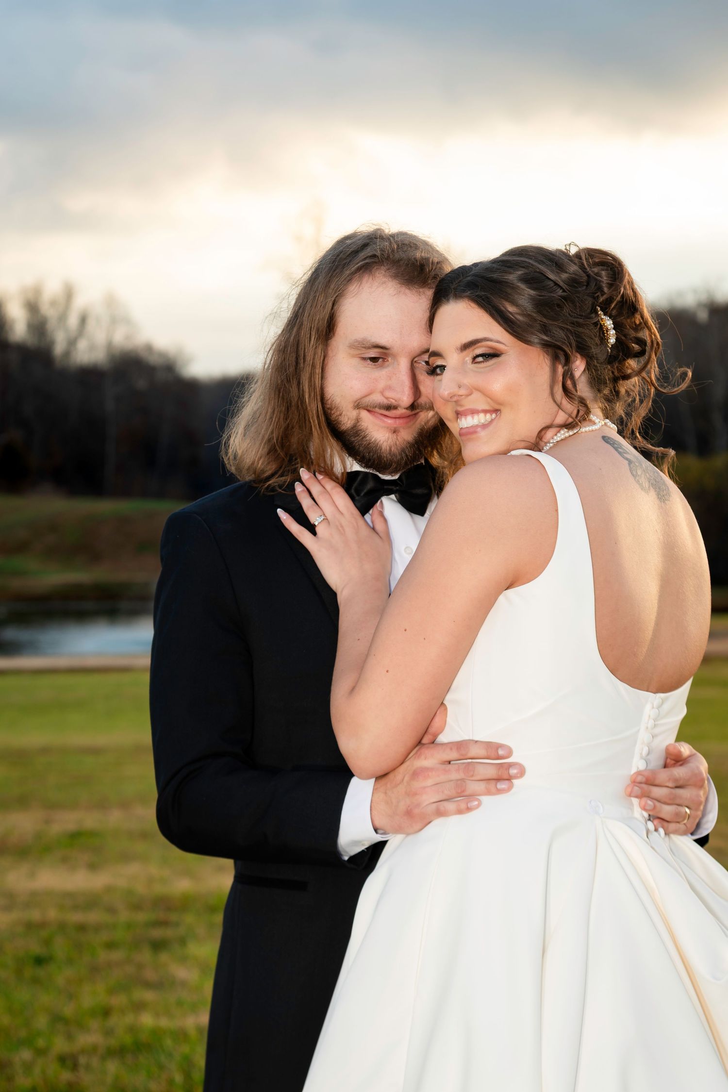 Groom embraces bride from behind as she smiles at camera during golden hour wedding portraits.