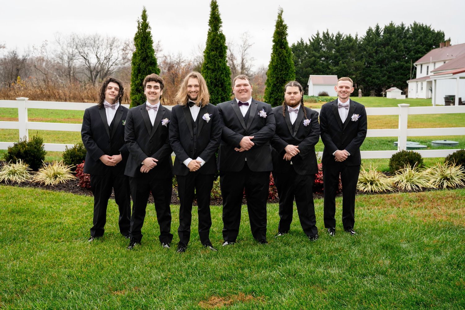 Groom and five groomsmen in black suits with bow ties standing by white fence at outdoor wedding venue.