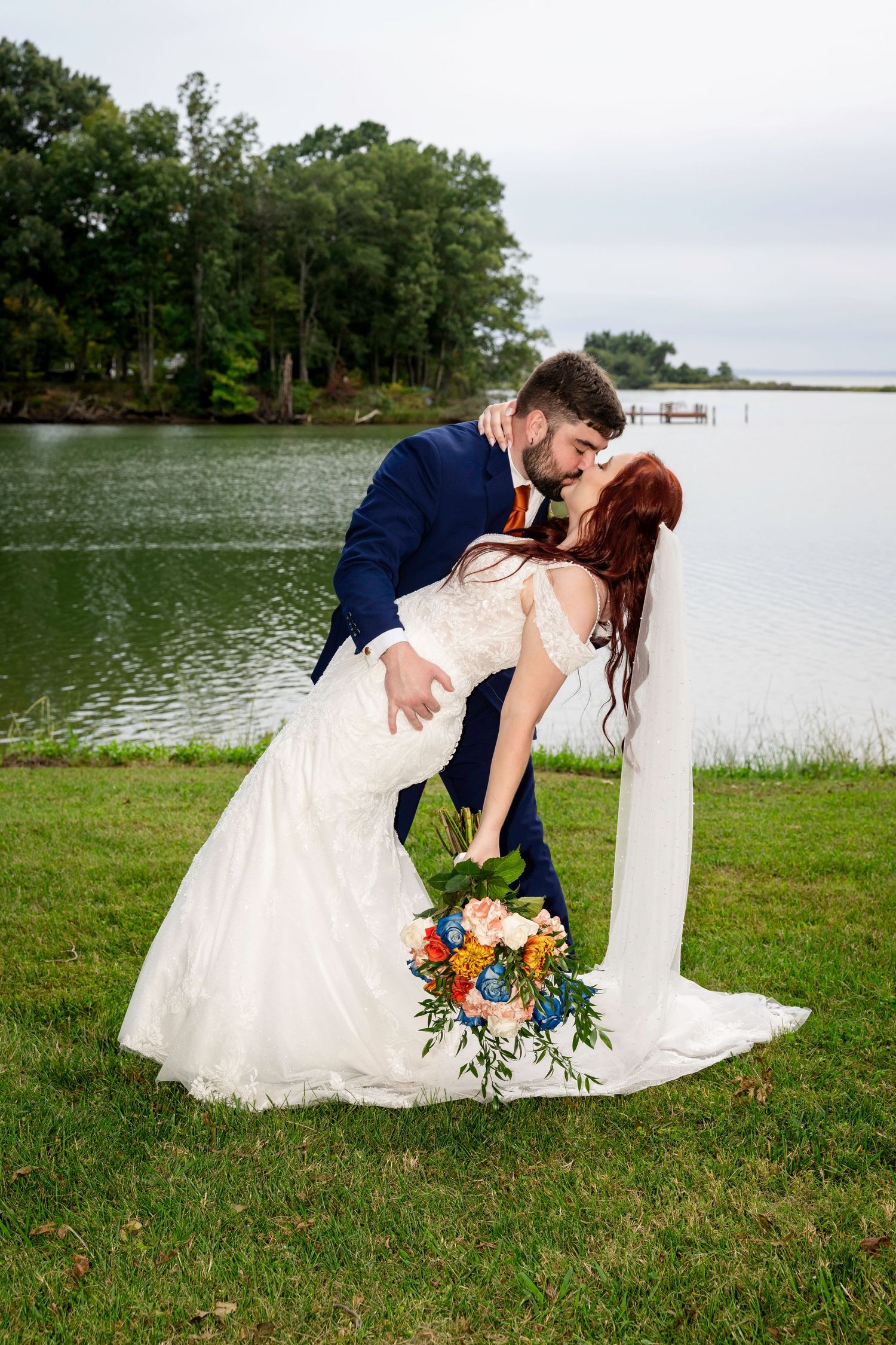 Groom dips bride for a kiss by lakeside, her white mermaid gown and colorful bouquet trailing on green grass.