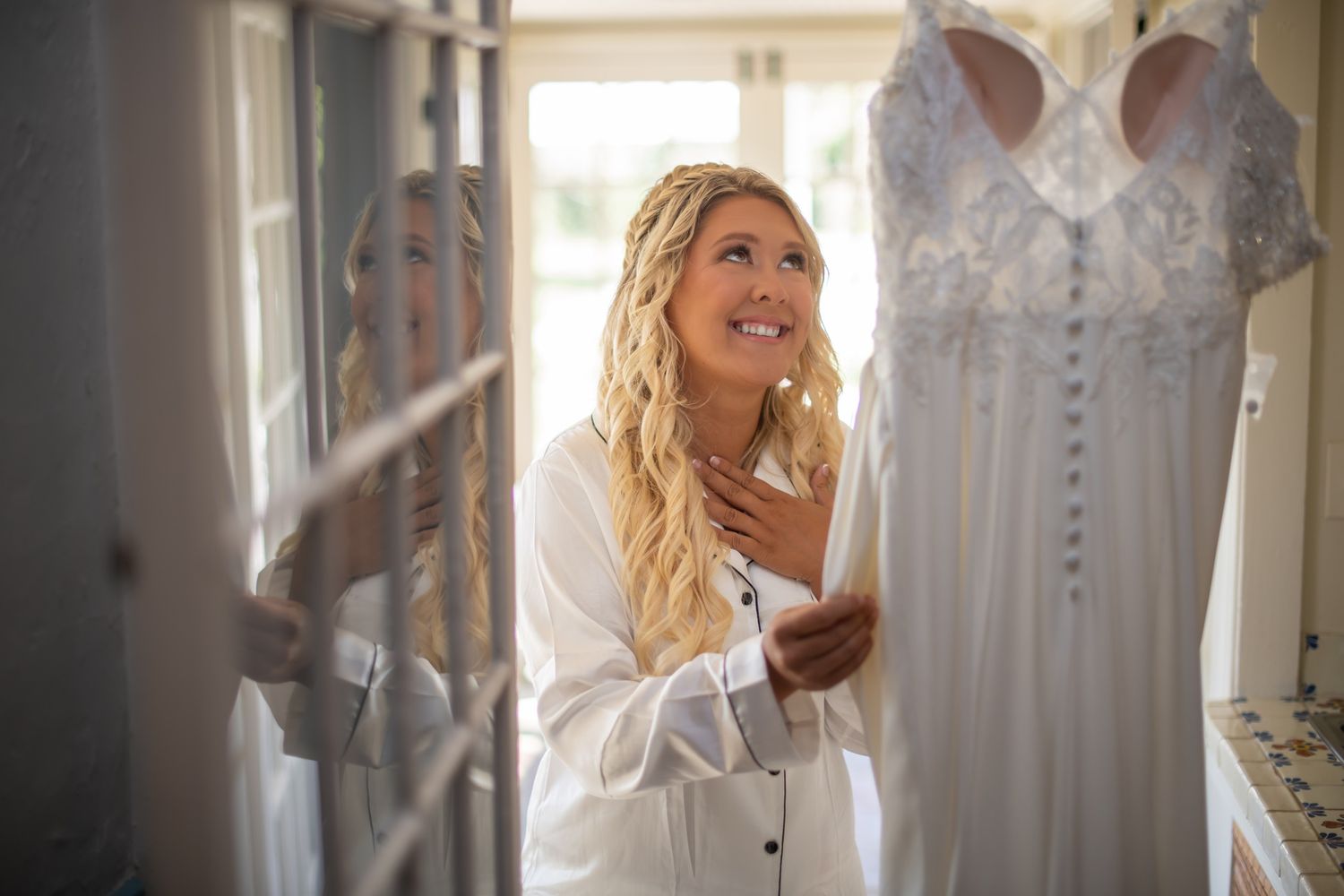 Smiling blonde bride in white robe admiring her lace wedding dress hanging in sunlit room.