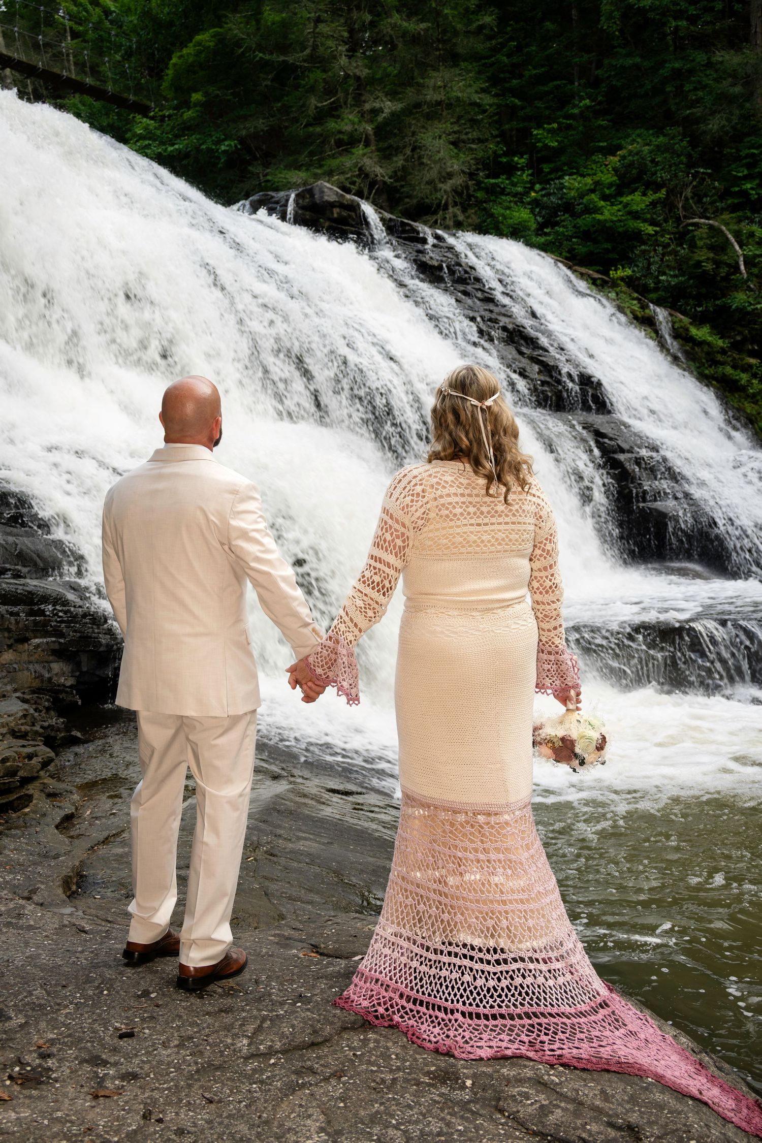 Bride and groom hold hands facing a cascading waterfall, bride wearing a crochet ombre wedding dress.