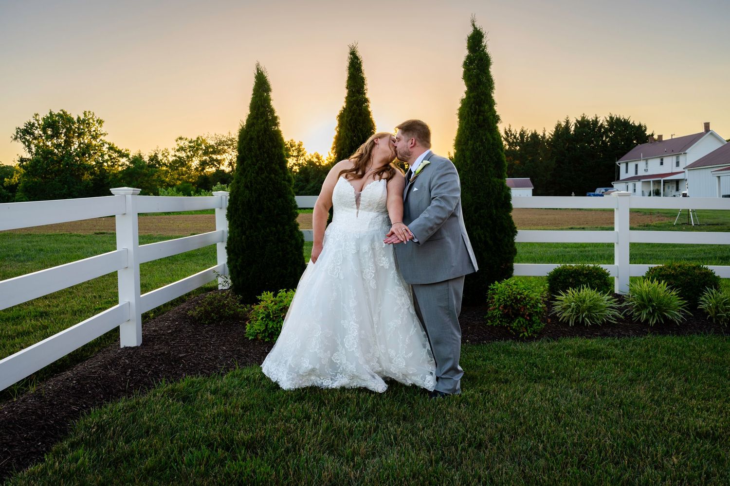 A bride and groom share a romantic kiss at sunset beside tall evergreen trees and a white fence at a farm venue.