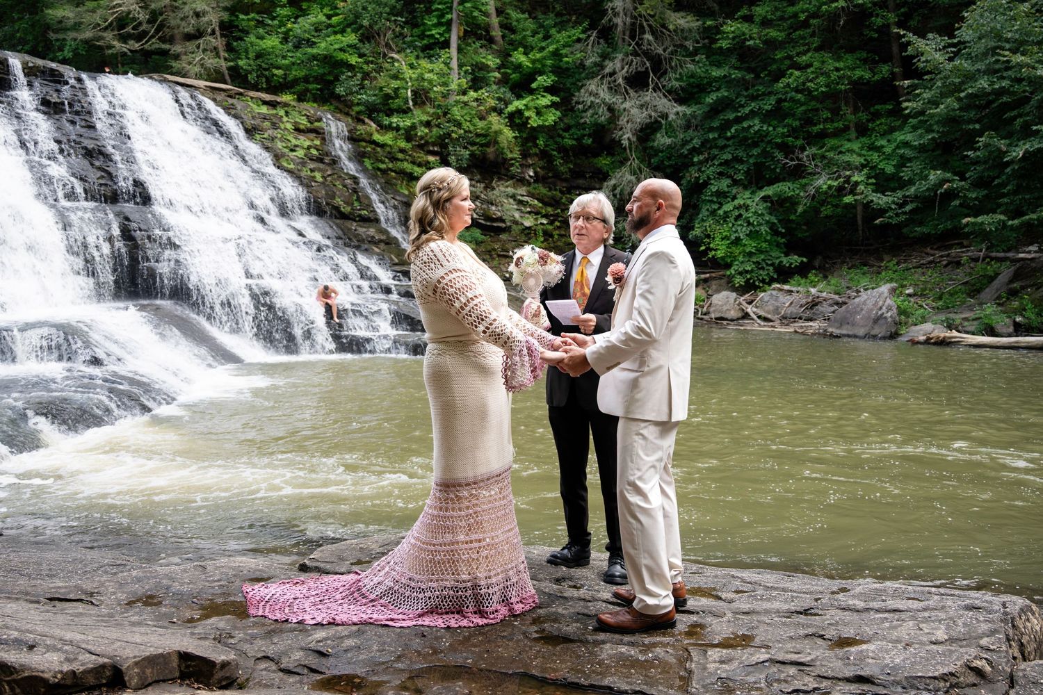 Couple exchanges vows at waterfall wedding ceremony outdoors, officiant holding flowers, rocky riverbank backdrop.