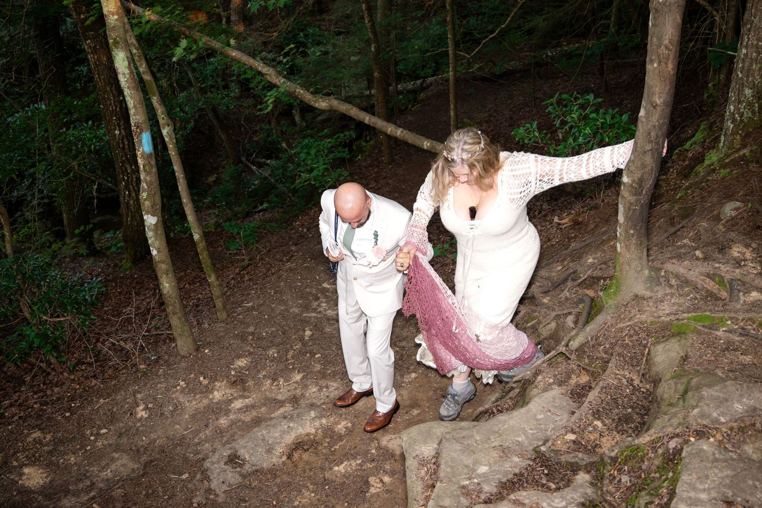 Bride and groom in wedding attire walking down stone steps in a lush forest setting.