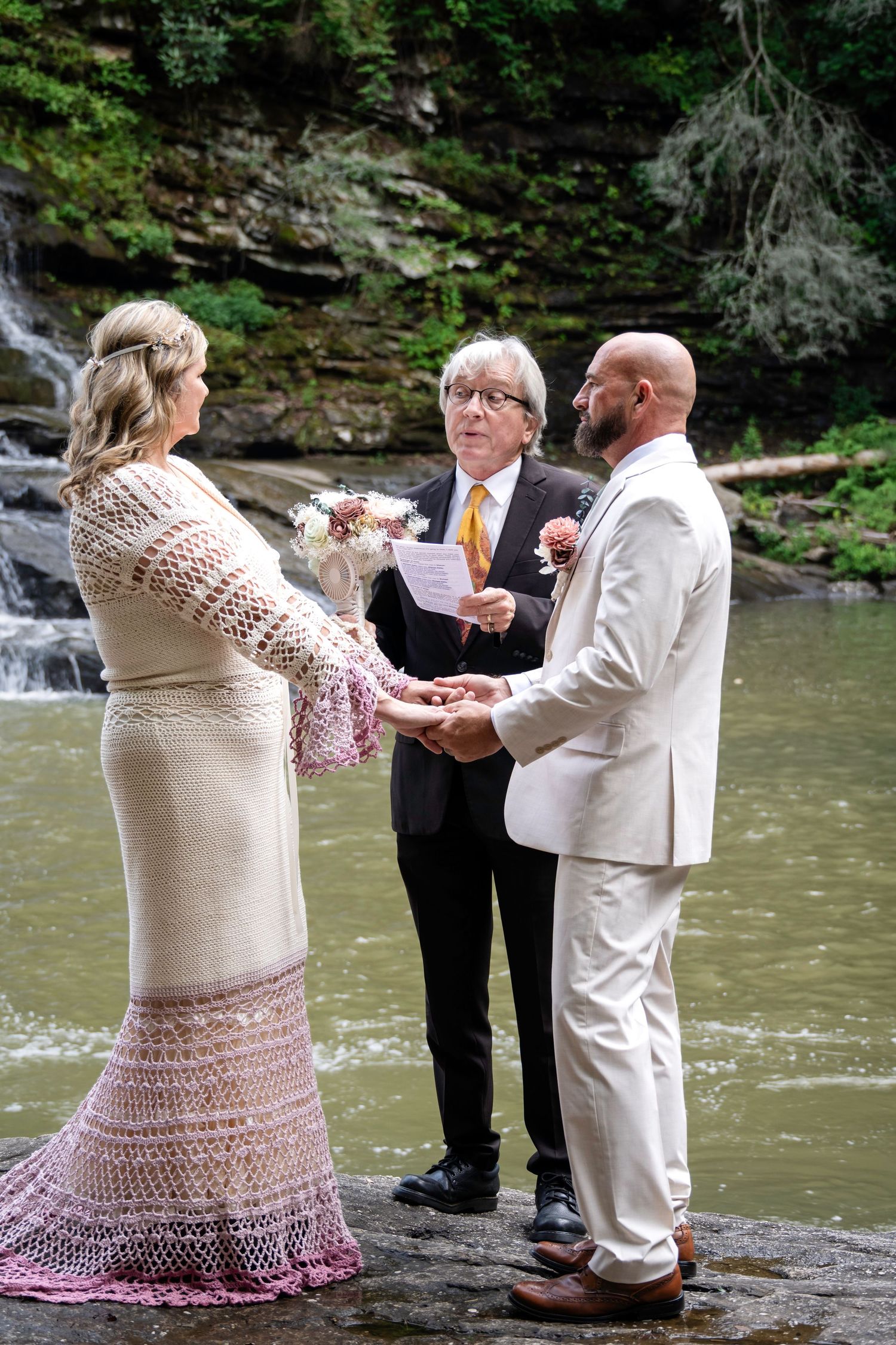 Couple exchanging vows at waterfall wedding ceremony with officiant holding papers, bride in lace dress, groom in tan suit.