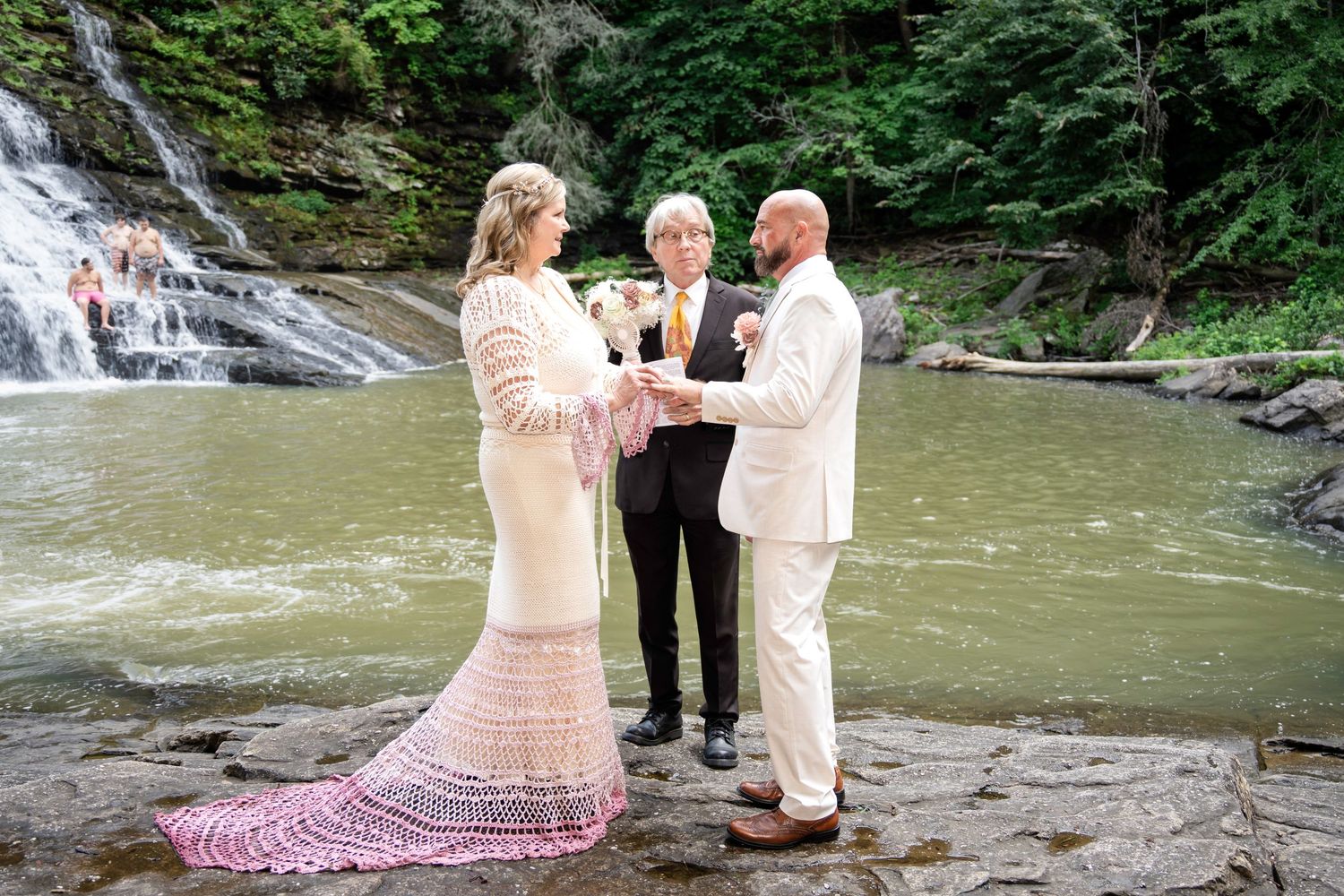 Couple exchanging vows at a waterfall ceremony, bride in pink lace gown, groom in white suit, officiant holding bouquet.