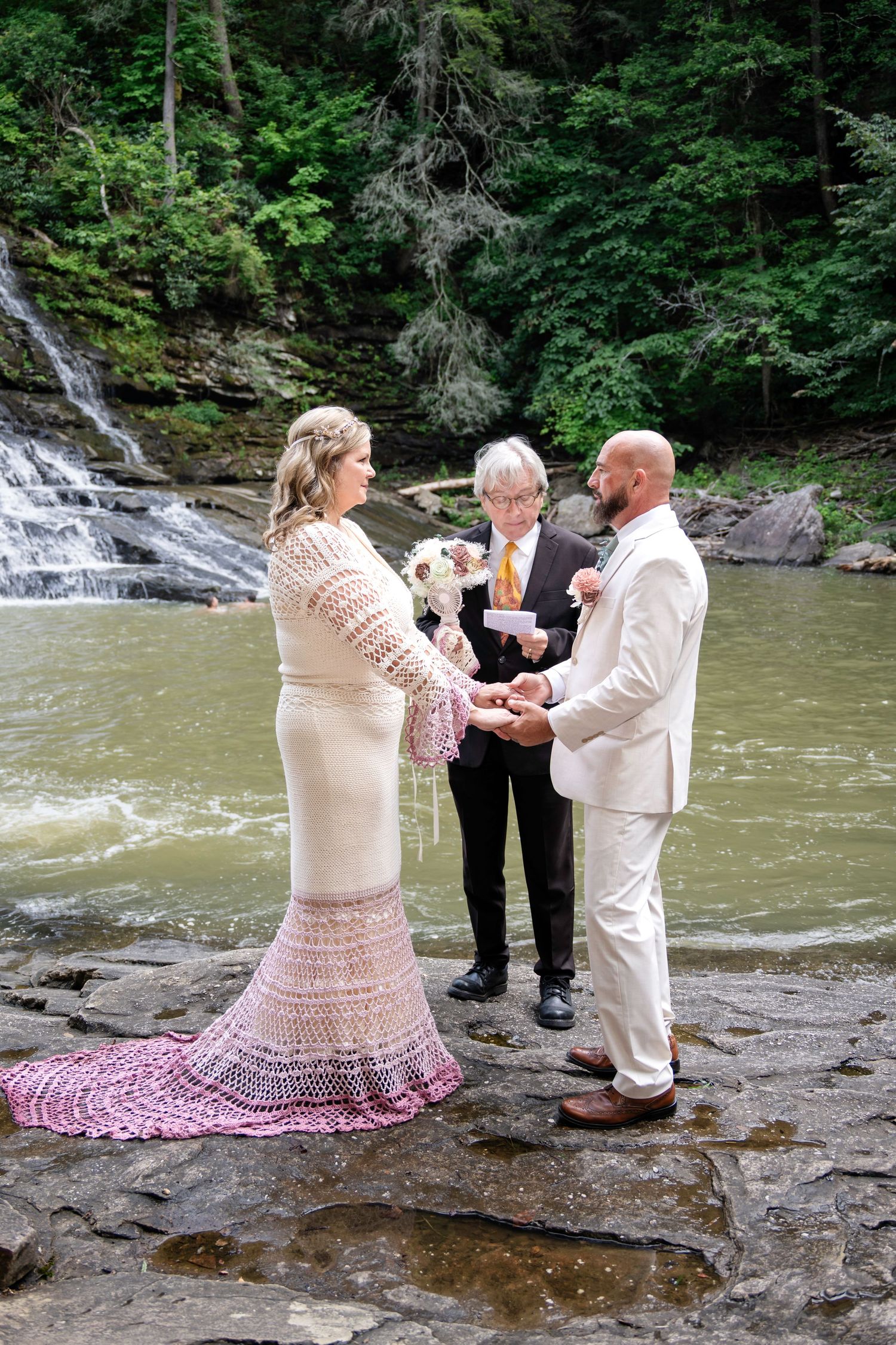 Couple exchanges vows beside a waterfall on rocky terrain, officiated by a minister in a lush forest setting.