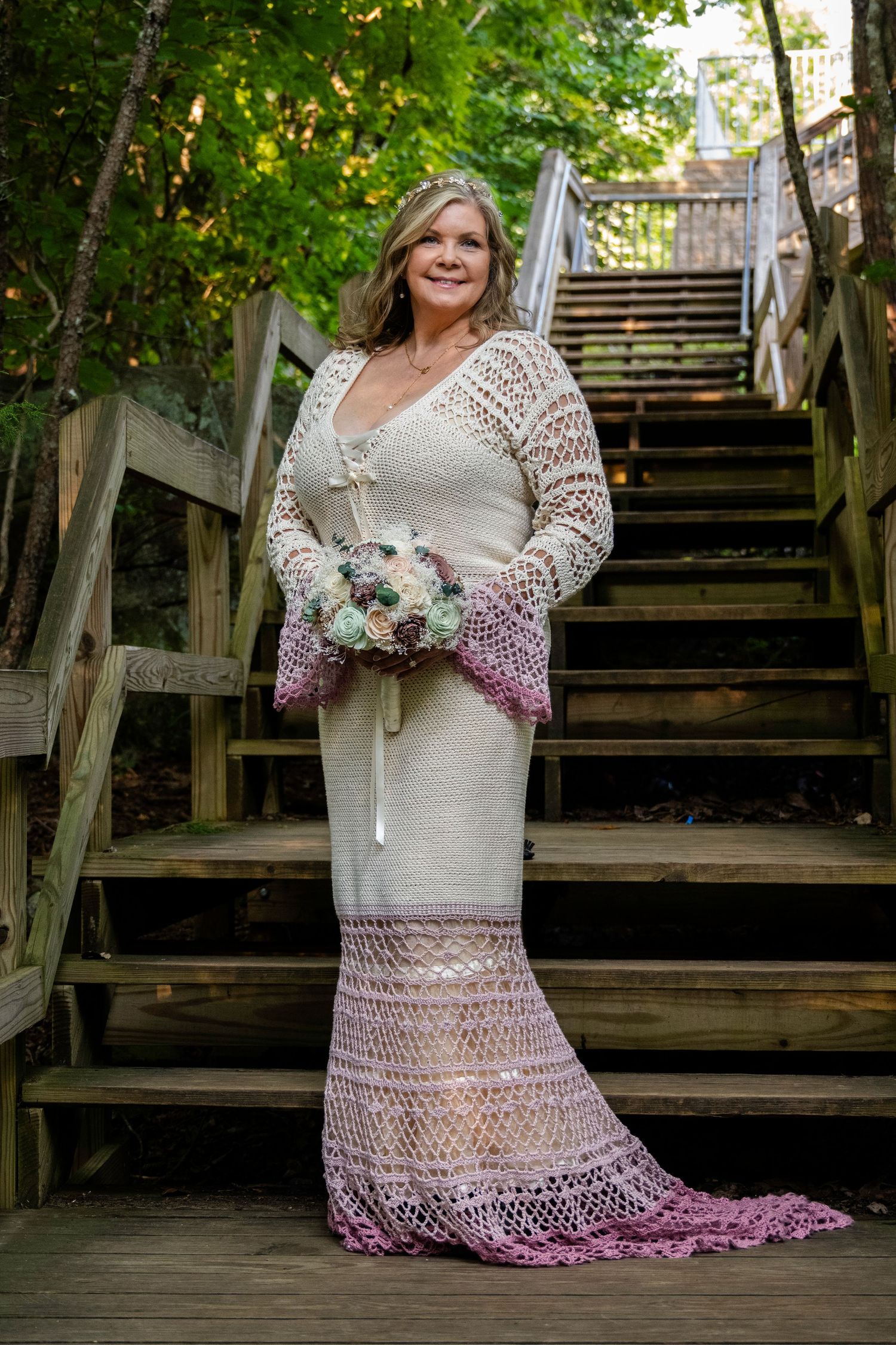 A bride in a white crochet wedding dress with pink trim holds a bouquet on wooden stairs outdoors.