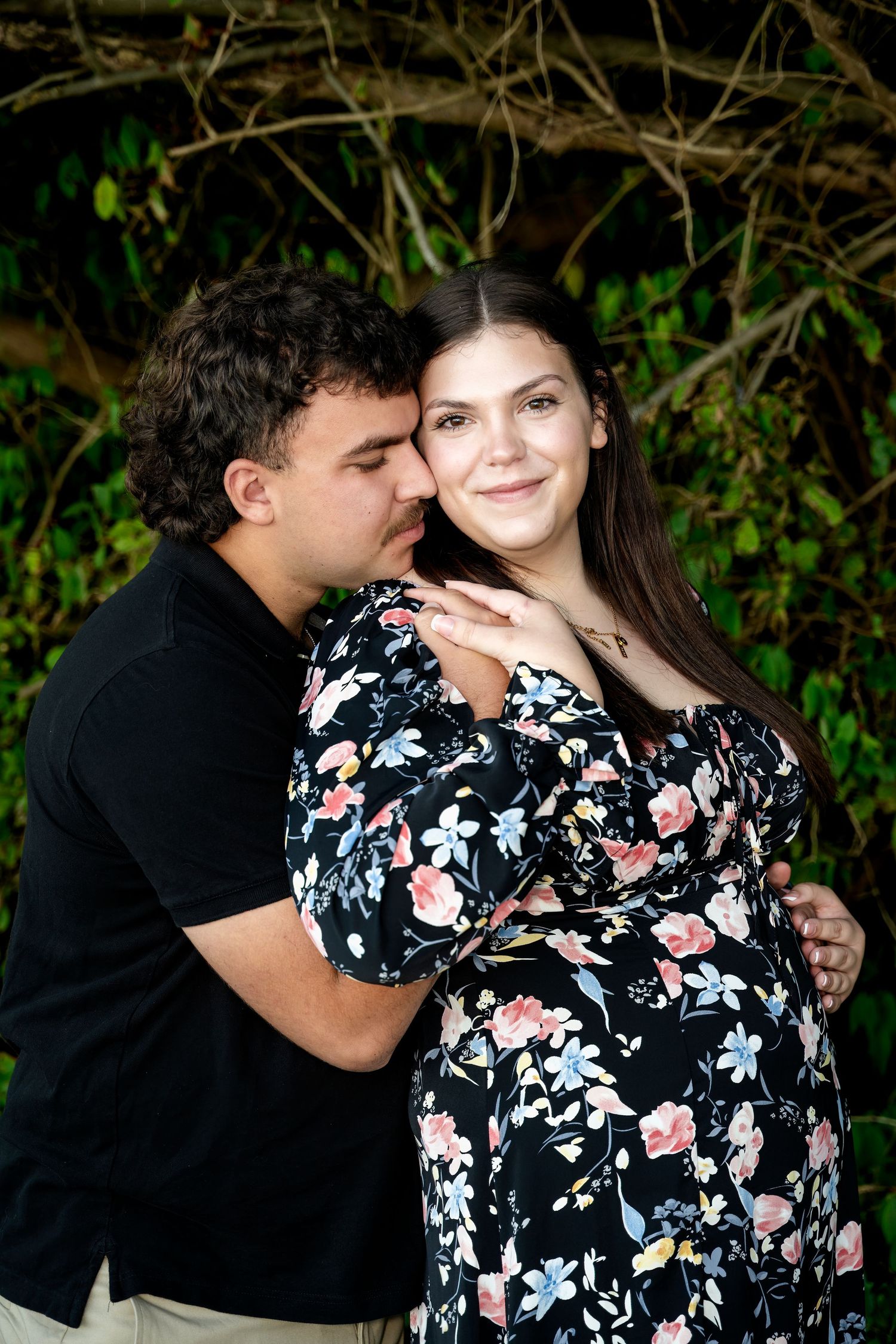 A man kisses a pregnant woman in a floral dress as she smiles, posing together in front of lush green foliage.