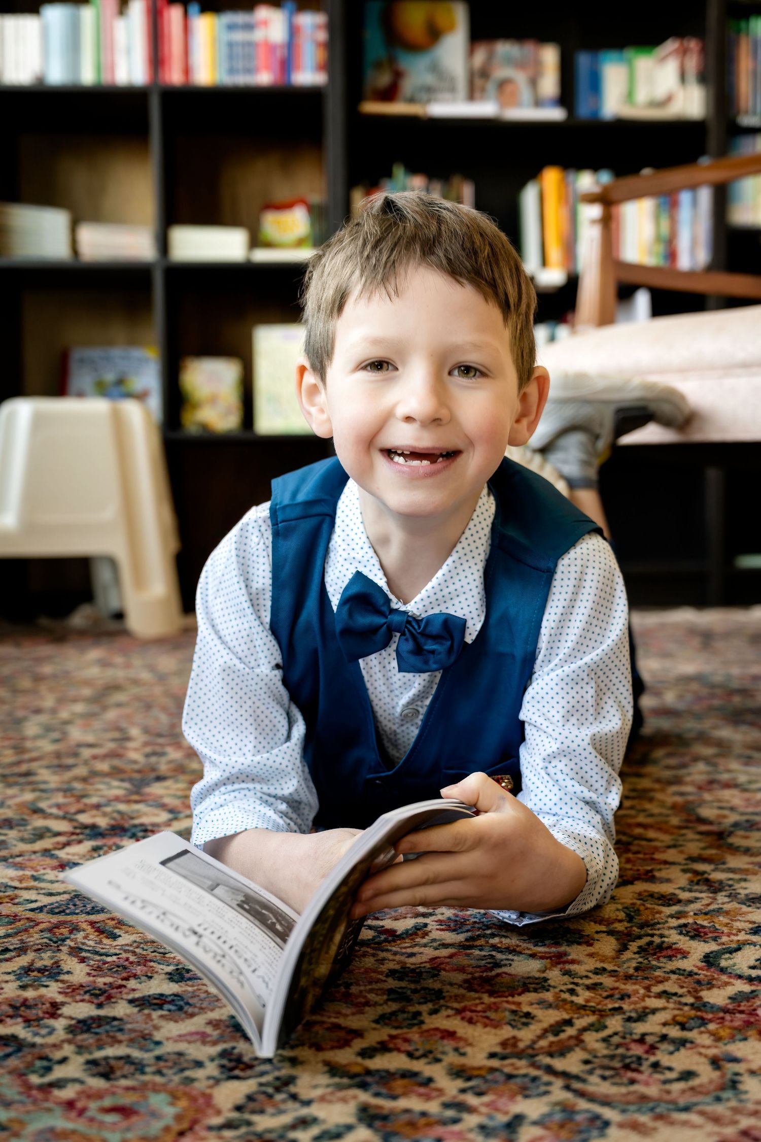Smiling young boy in blue vest and bow tie lying on carpet reading a magazine in a library.