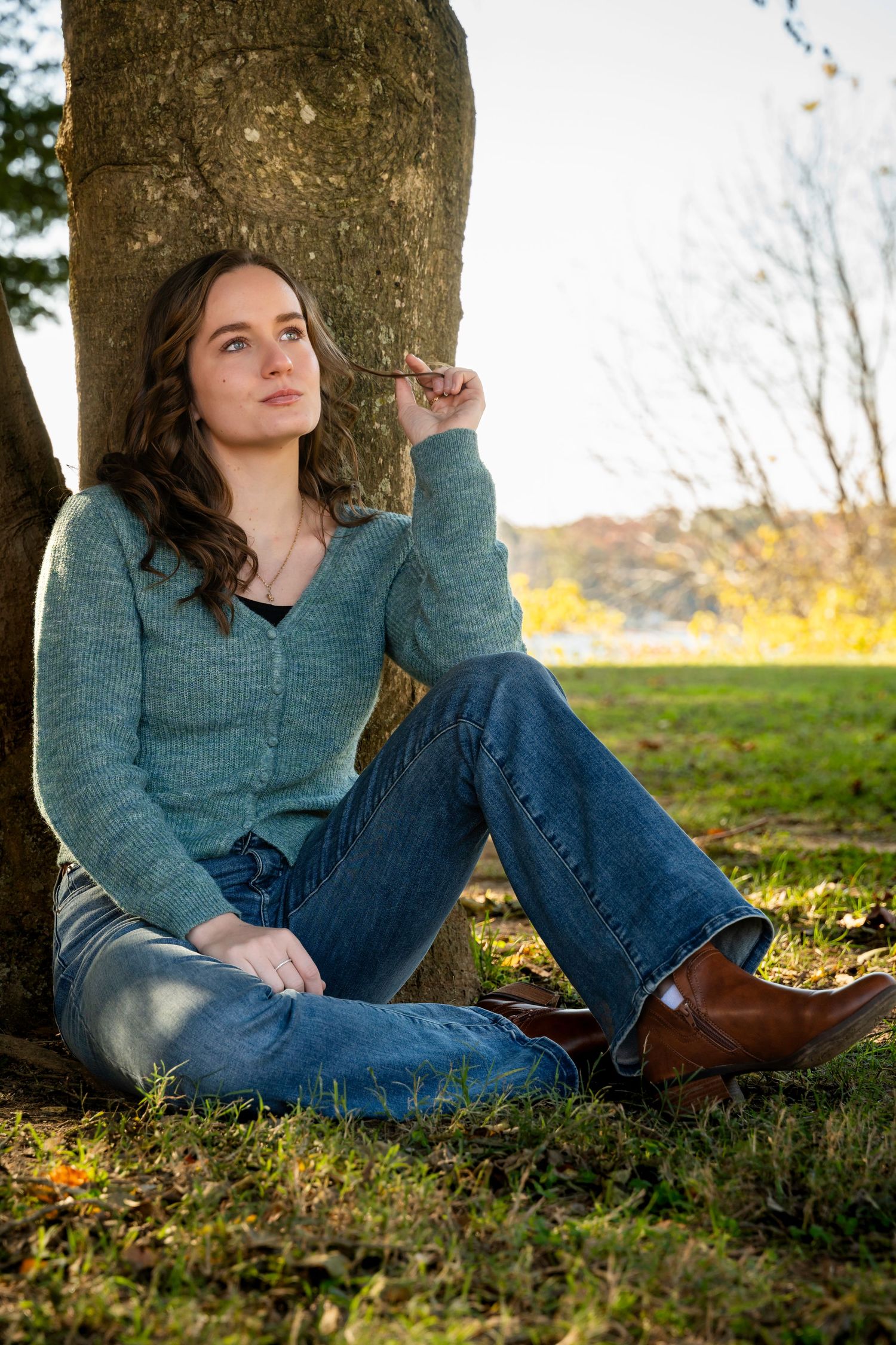 Young woman in teal sweater and jeans sits against tree trunk in autumn park, gazing thoughtfully upward.