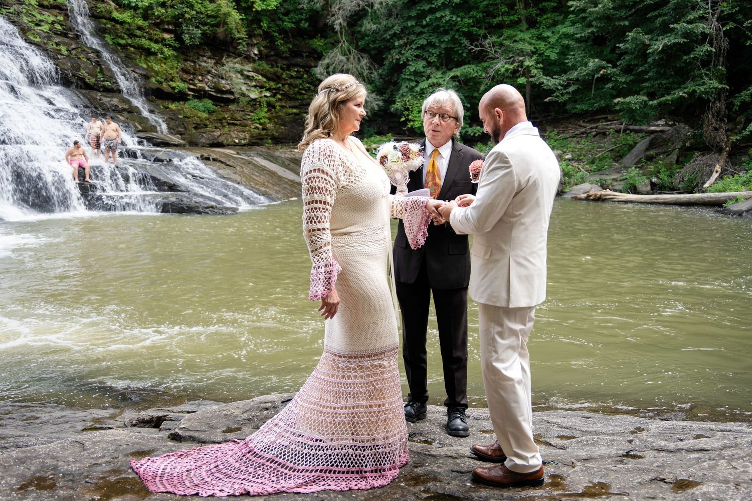 Outdoor wedding ceremony by a waterfall, bride in lace gown, groom in white suit, officiant joining hands.