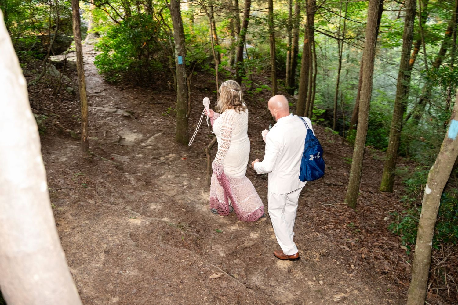 Couple in white wedding attire hiking a forest trail, bride holding bouquet, groom wearing blue backpack.