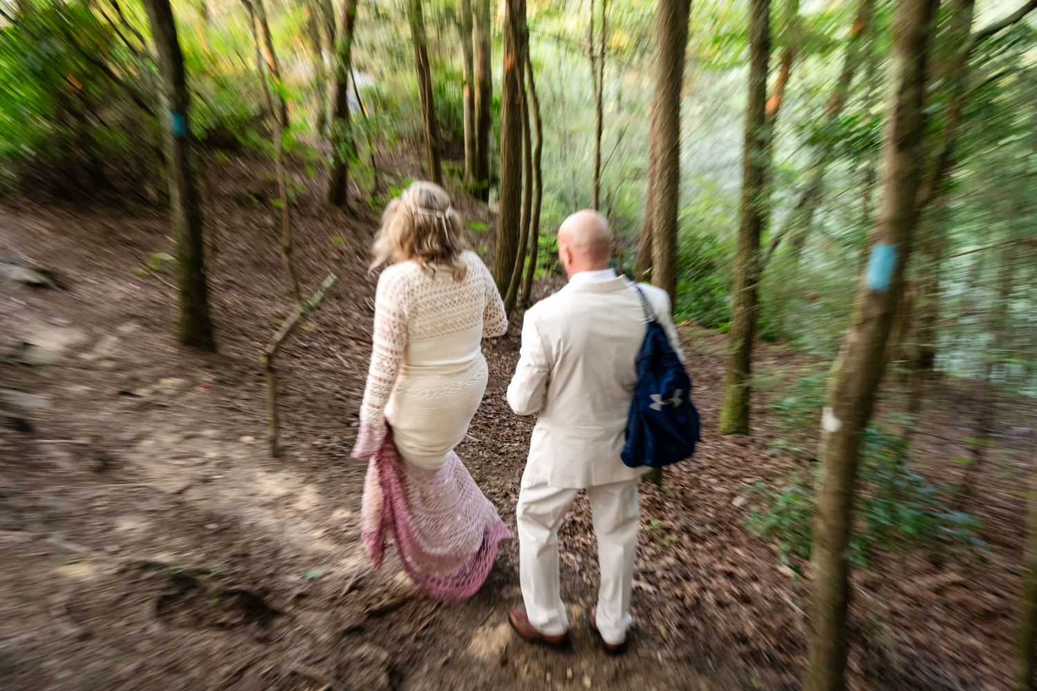 Couple walking hand-in-hand through a lush forest trail, woman in white lace dress, man in beige suit with blue backpack.