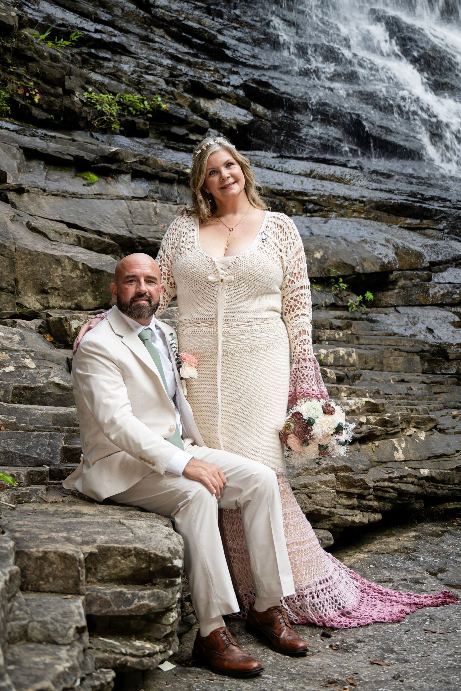 Bride and groom pose on stone steps before a waterfall, she in a crochet gown, he in a tan suit.