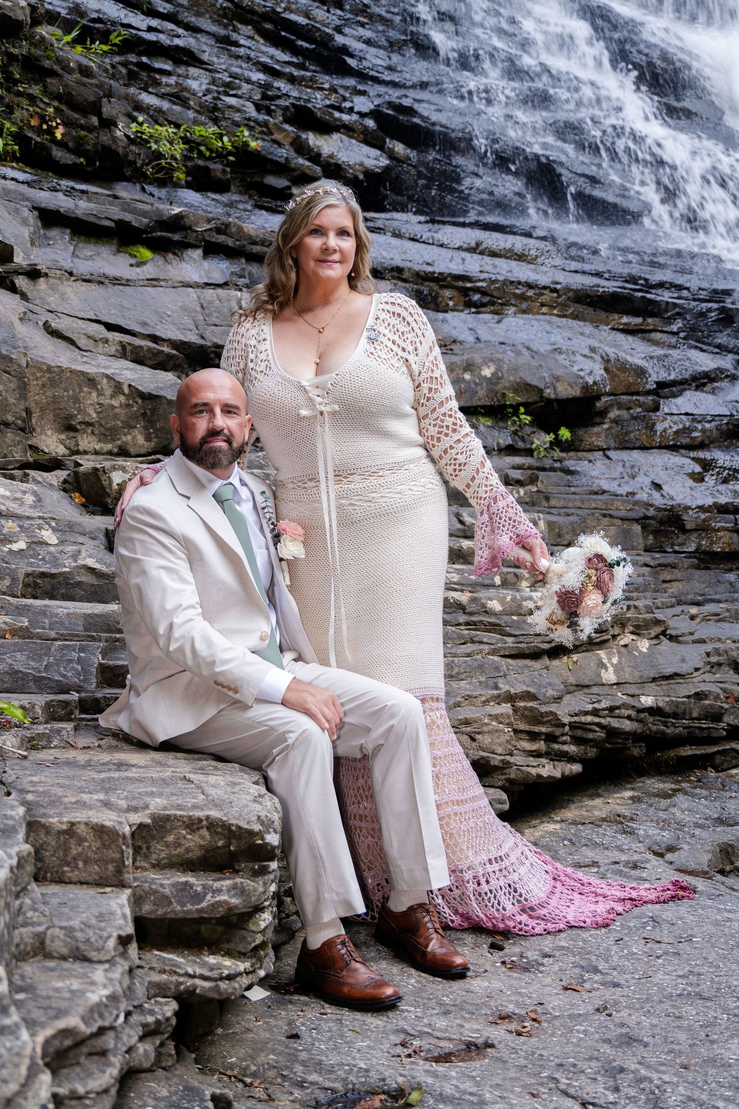Bride and groom pose on rocky steps before a cascading waterfall, bride wearing crochet wedding dress with pink ombre train.