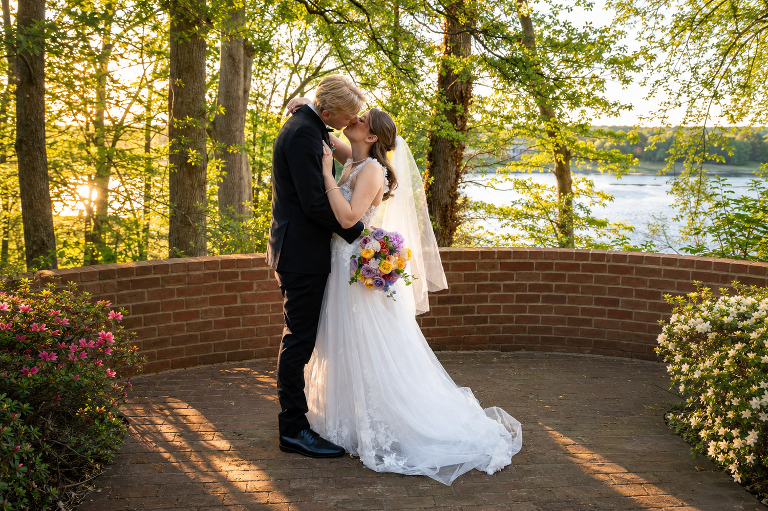 Bride and groom share a romantic kiss on a brick terrace surrounded by lush trees and a serene lake at sunset.