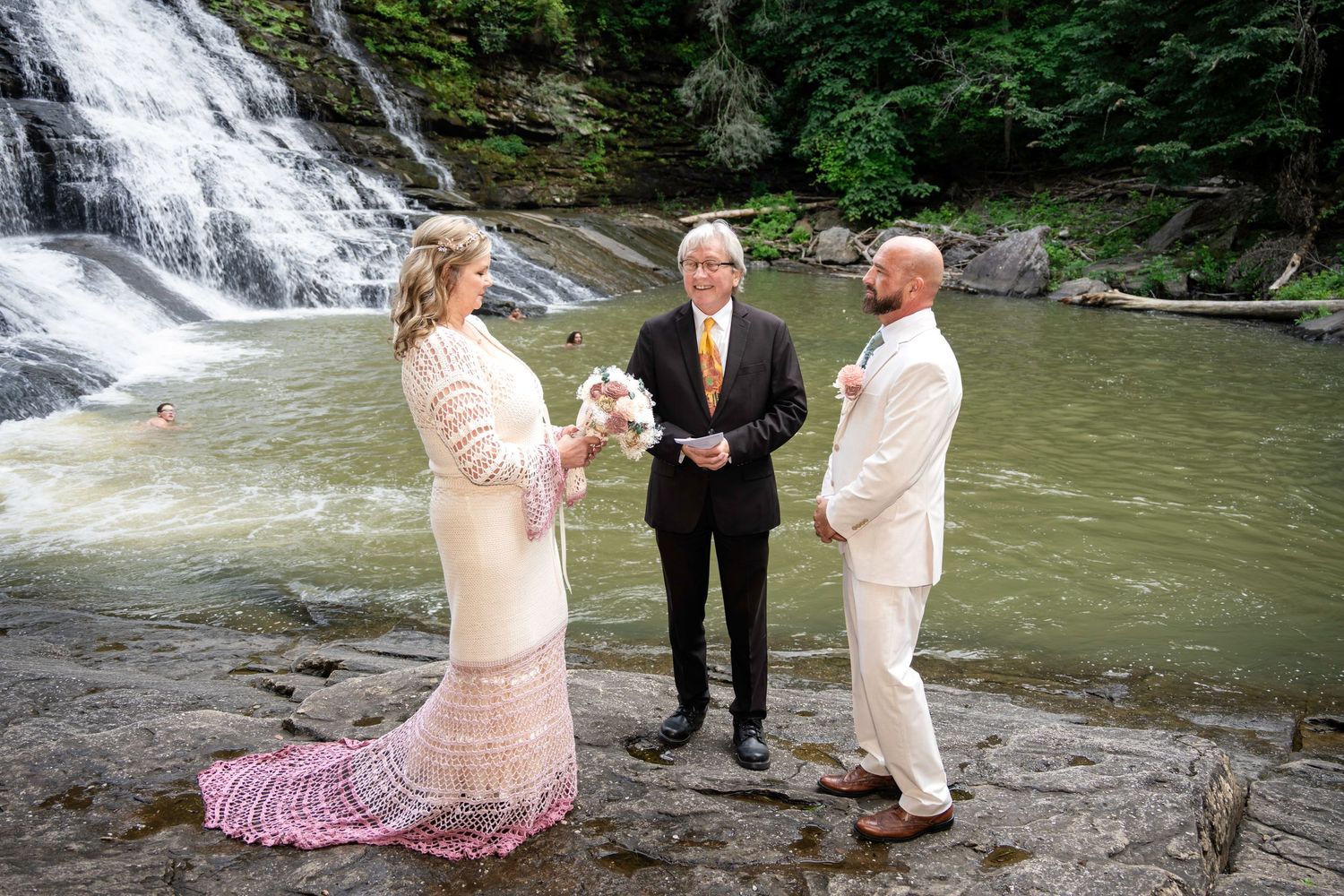 Couple exchanges vows with officiant beside a waterfall, bride in lace gown, groom in white shirt, lush green backdrop.