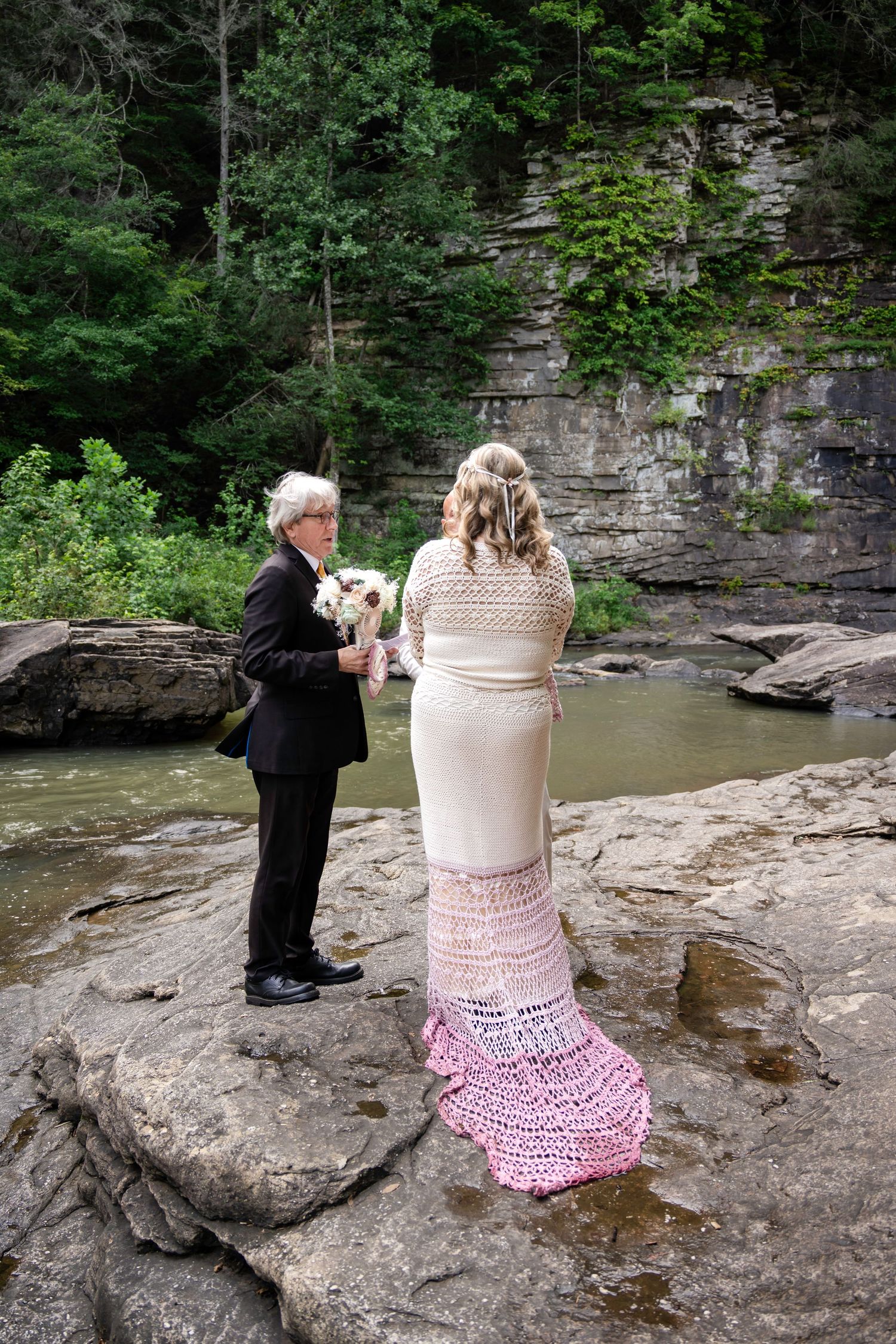 Couple exchanging vows during an outdoor wedding ceremony on rocky riverbank surrounded by lush forest and stone cliffs.