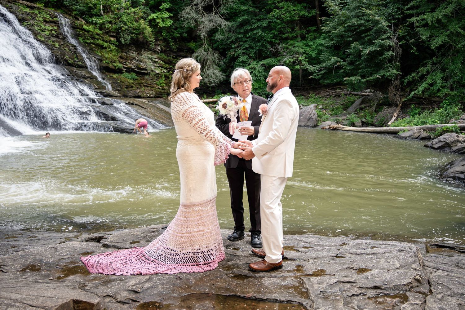Couple exchanging vows at a waterfall wedding ceremony on rocks, officiant holding flowers, lush green forest backdrop.