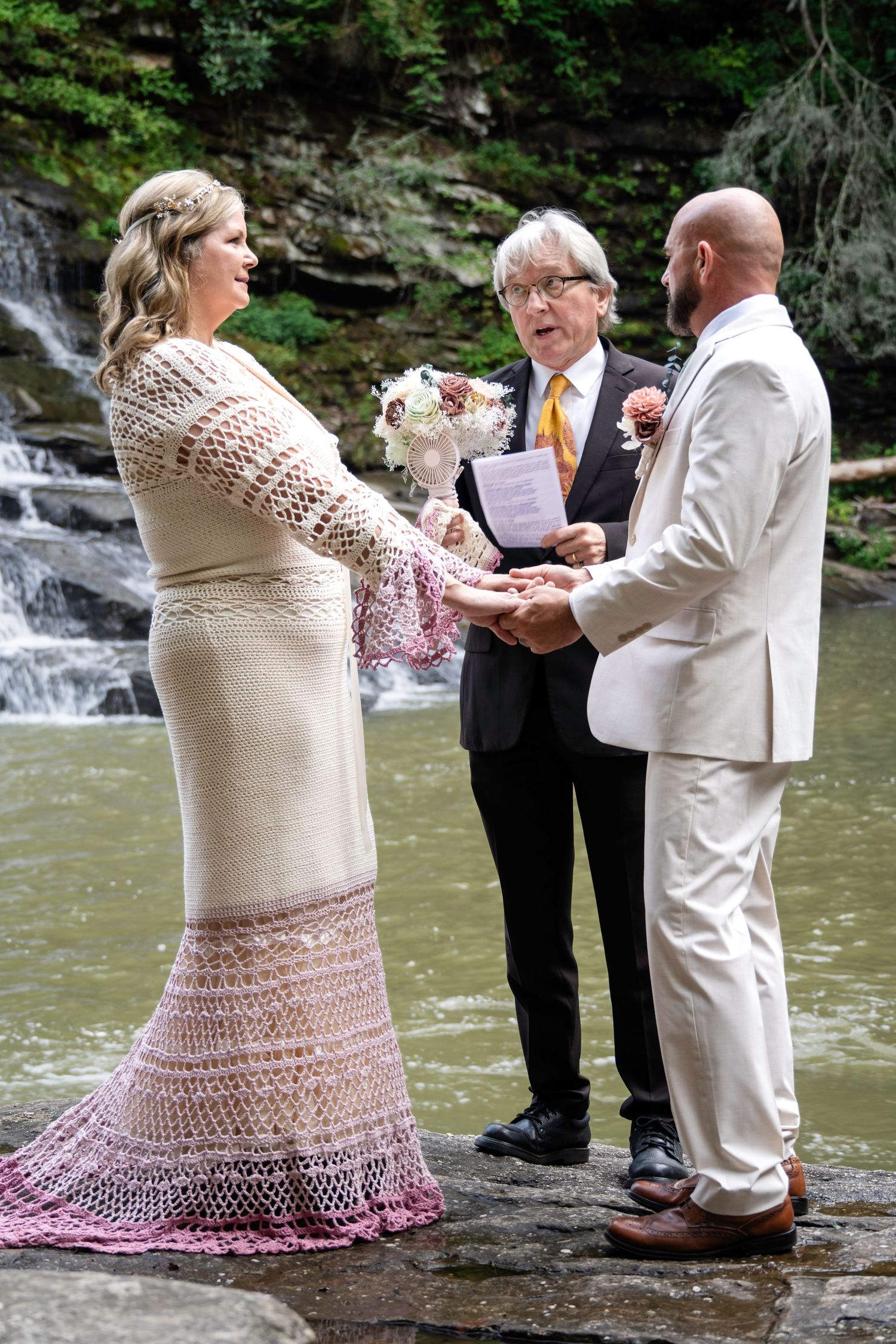 Couple exchanges vows at waterfall wedding ceremony, bride in crochet gown, groom in tan suit, officiant in black.