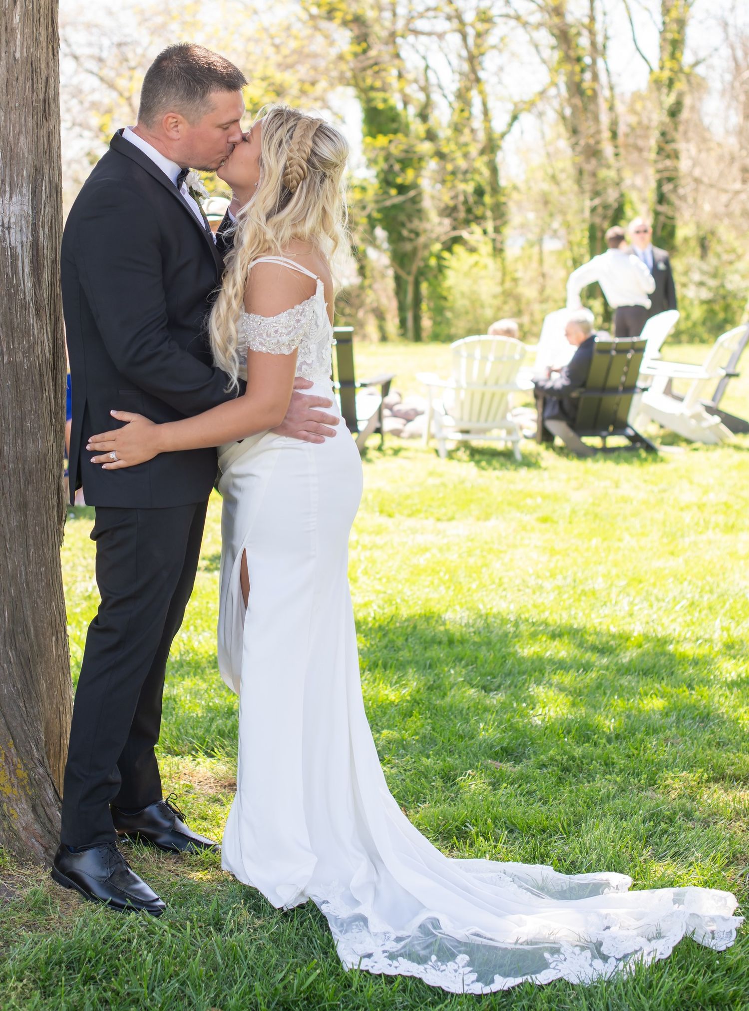 Bride and groom sharing romantic kiss beside tree in sunny garden, bride in off-shoulder lace gown. - Lusby Maryland