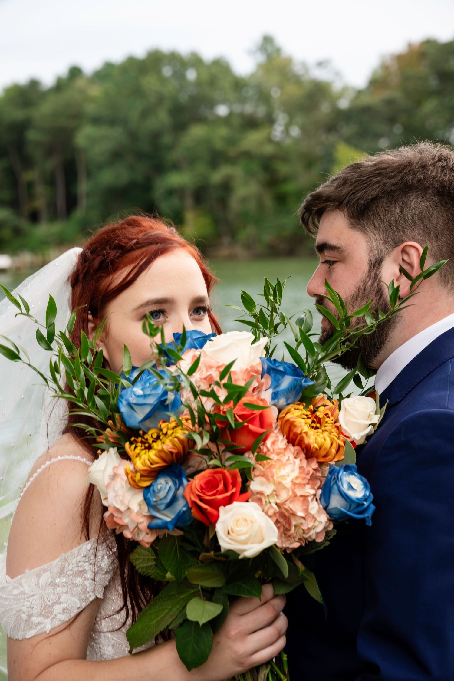 Bride and groom peek over a colorful bouquet of blue, orange, and peach roses in a close romantic portrait.