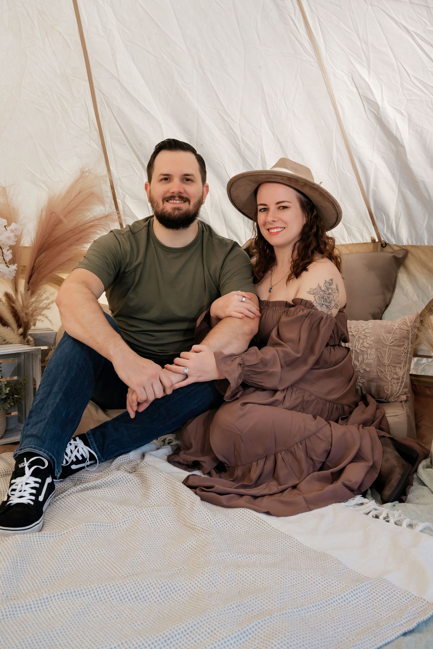 A couple sits together inside a boho-styled tent, smiling, holding hands, surrounded by pampas grass decor.