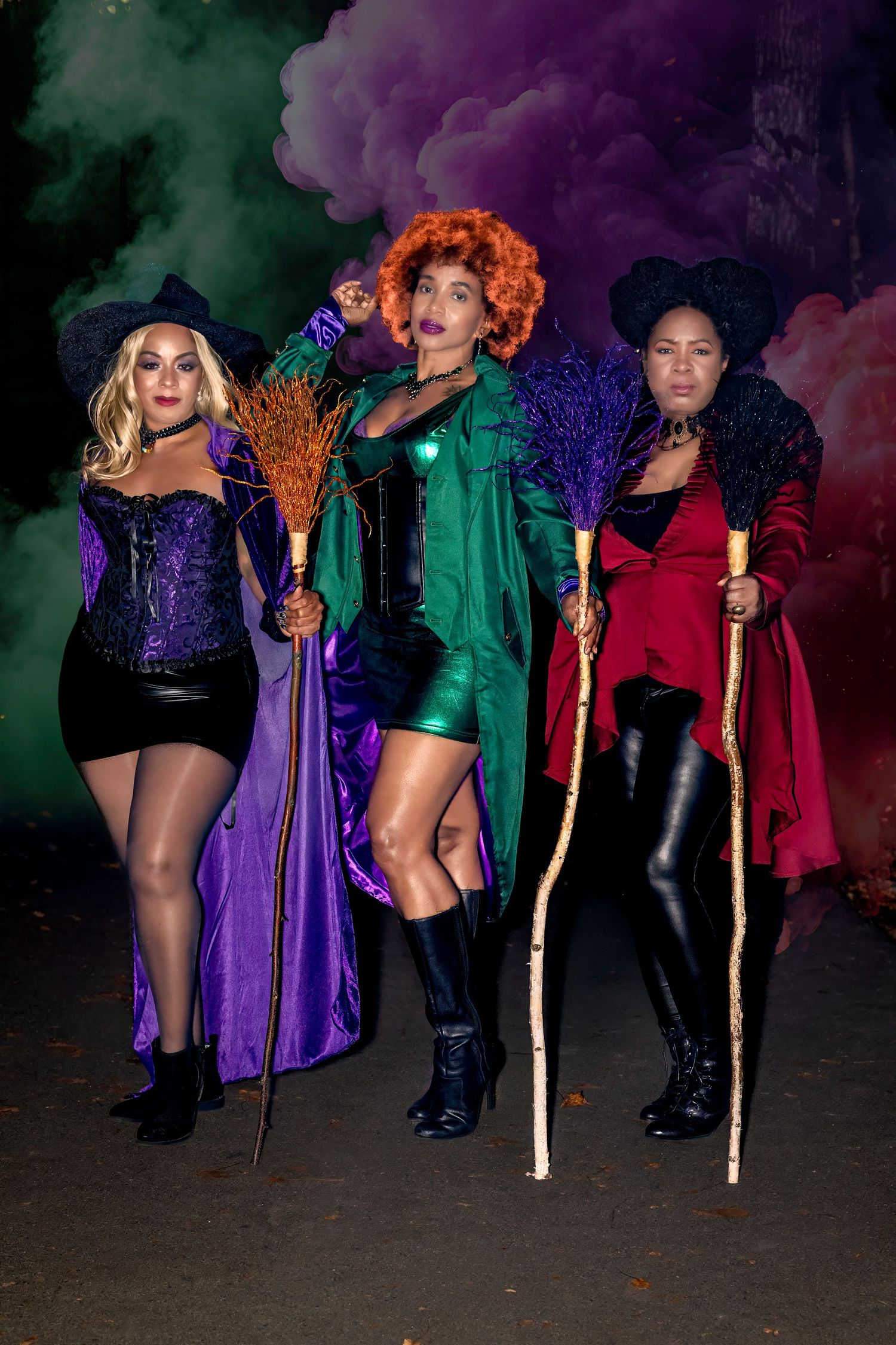 Three women in colorful witch Halloween costumes pose with brooms and staffs against dramatic green and purple smoke.