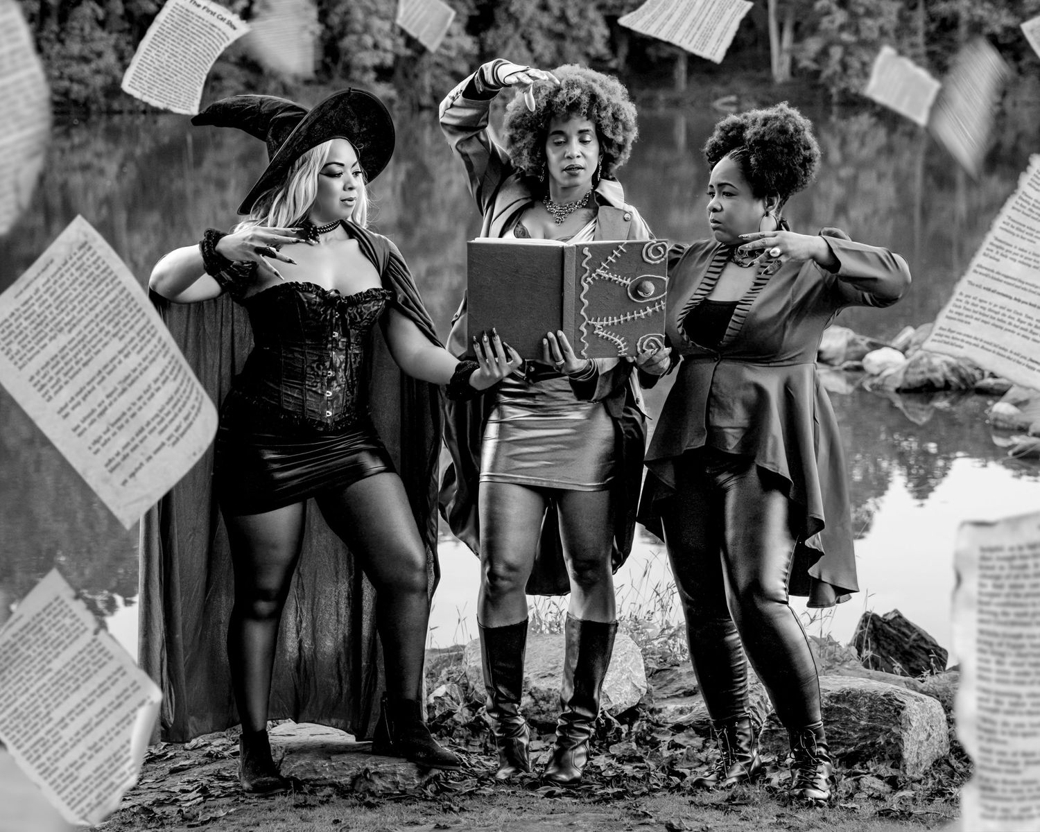 Three women in witch costumes pose dramatically outdoors with flying pages in black and white photo.