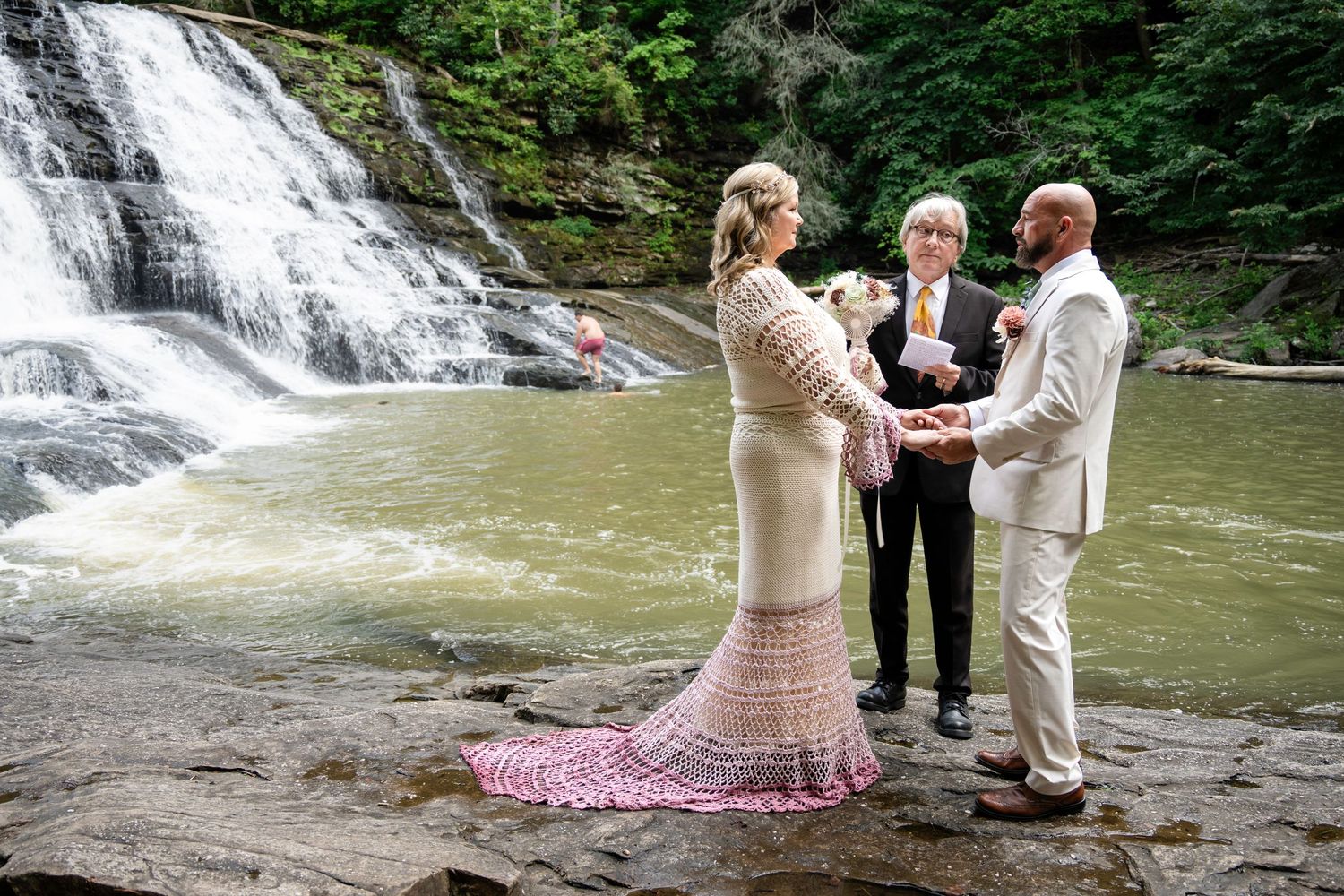 Couple exchanging vows at a waterfall wedding ceremony outdoors, officiant holding book, bride in lace gown, groom in white suit.